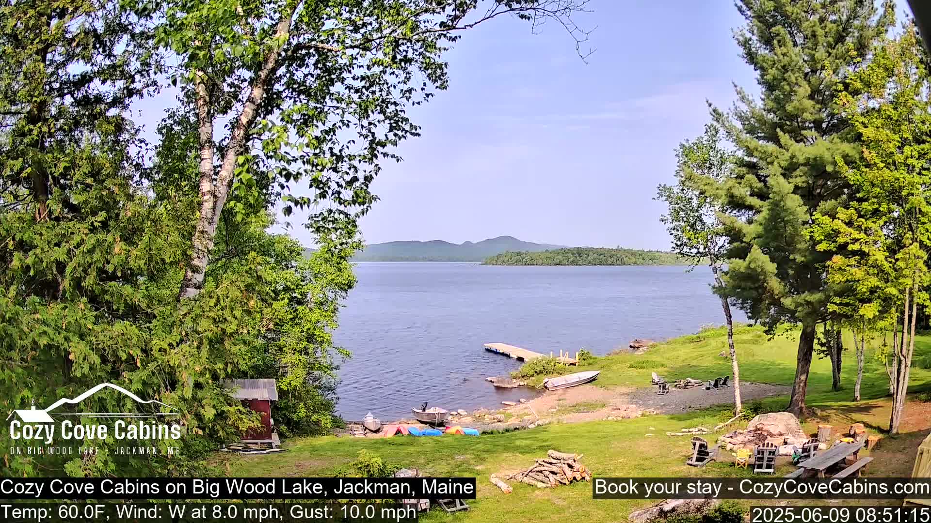 A lakeside view on a partly sunny day with a gentle westerly wind shows several boats pulled ashore near a small cabin and picnic table.