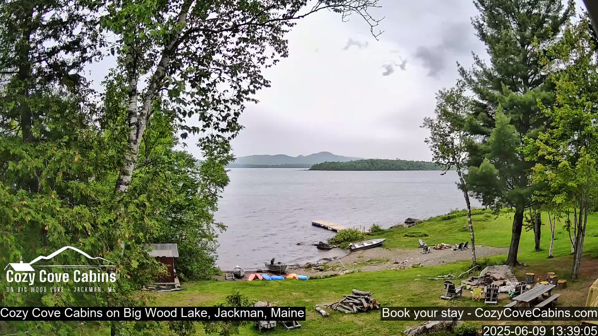 A lakeside view shows a calm lake under an overcast sky, with boats on a sandy shore, surrounded by lush green trees and a picnic area.