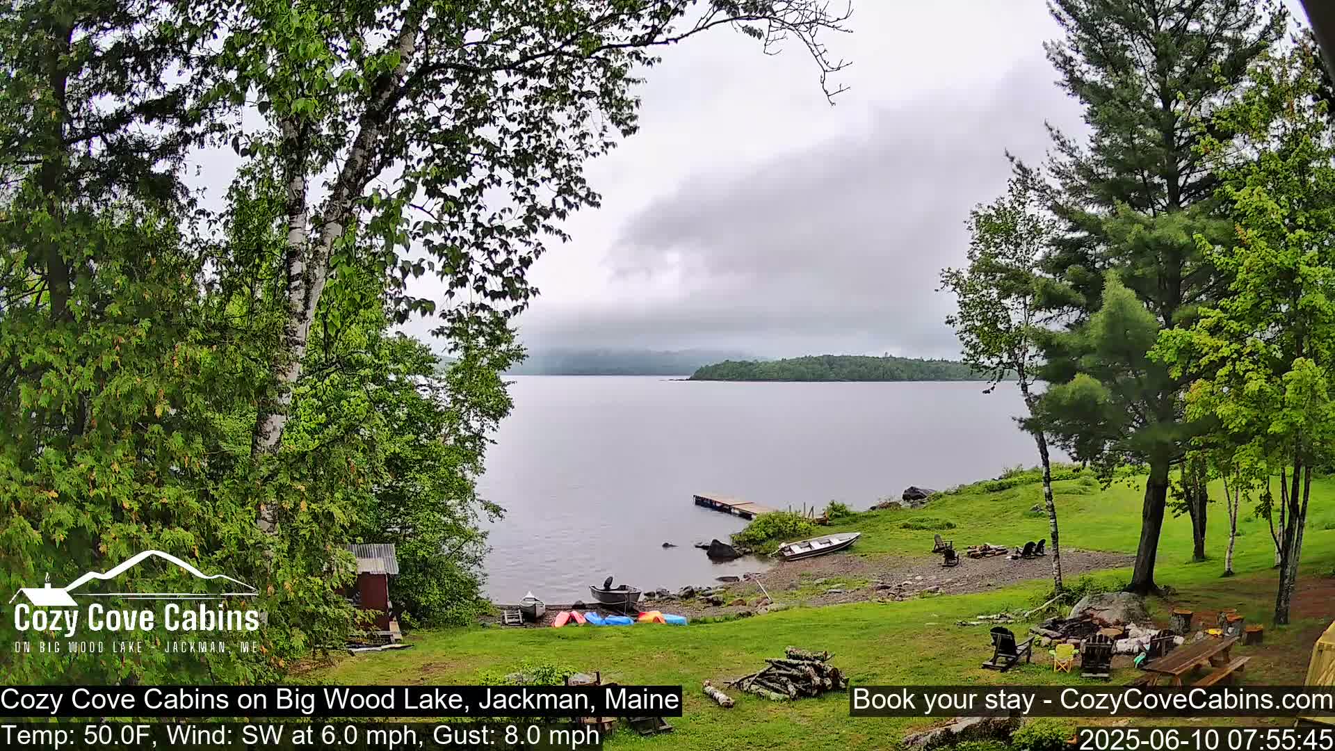 A lakeside view on an overcast day shows several boats pulled ashore near a small dock, with green grass, trees, and a campfire setup in the foreground.