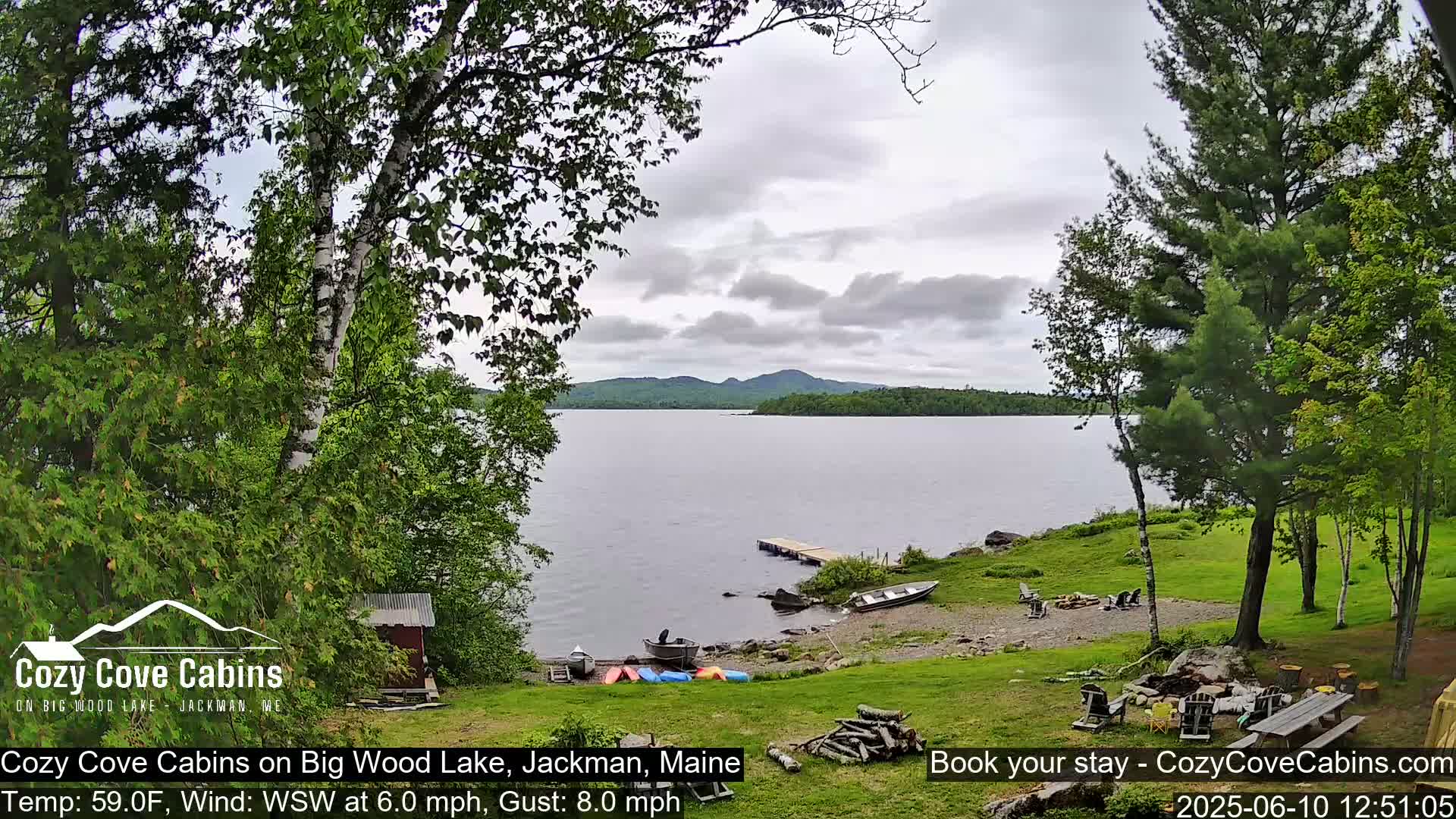 A tranquil lake scene under a cloudy sky shows a grassy shoreline with a few boats and outdoor furniture, surrounded by lush green trees.