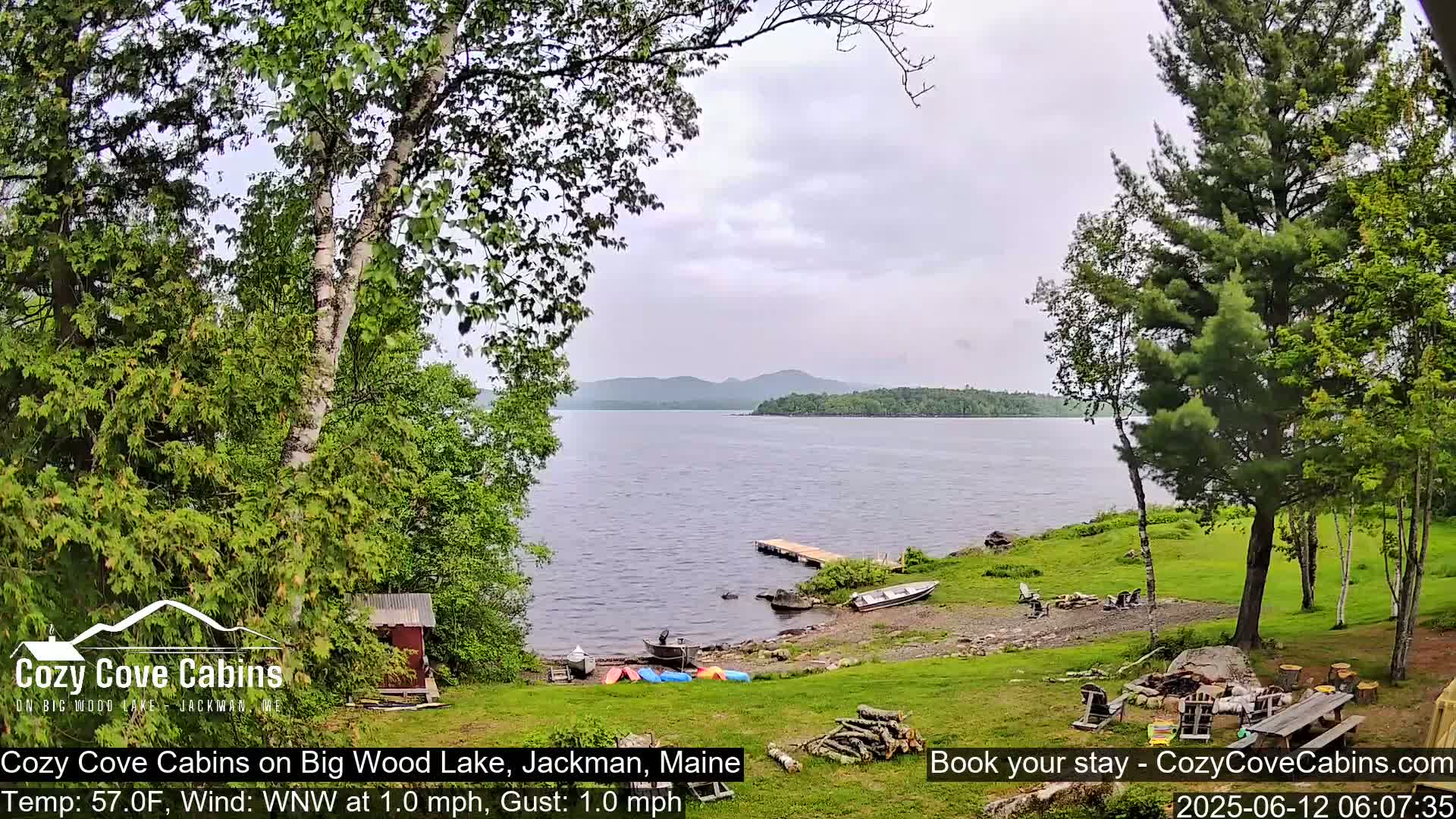 A lakeside property with several boats and kayaks on a grassy shoreline, under an overcast sky.