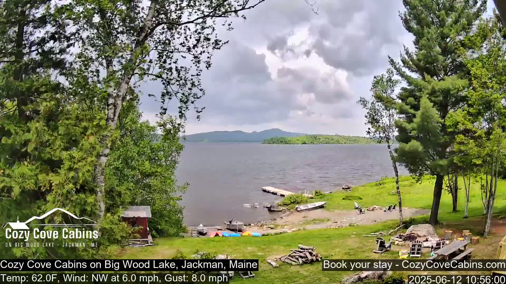 A lakeside view shows a calm lake under a partly cloudy sky, with several boats docked near a grassy shore where outdoor furniture is set up.