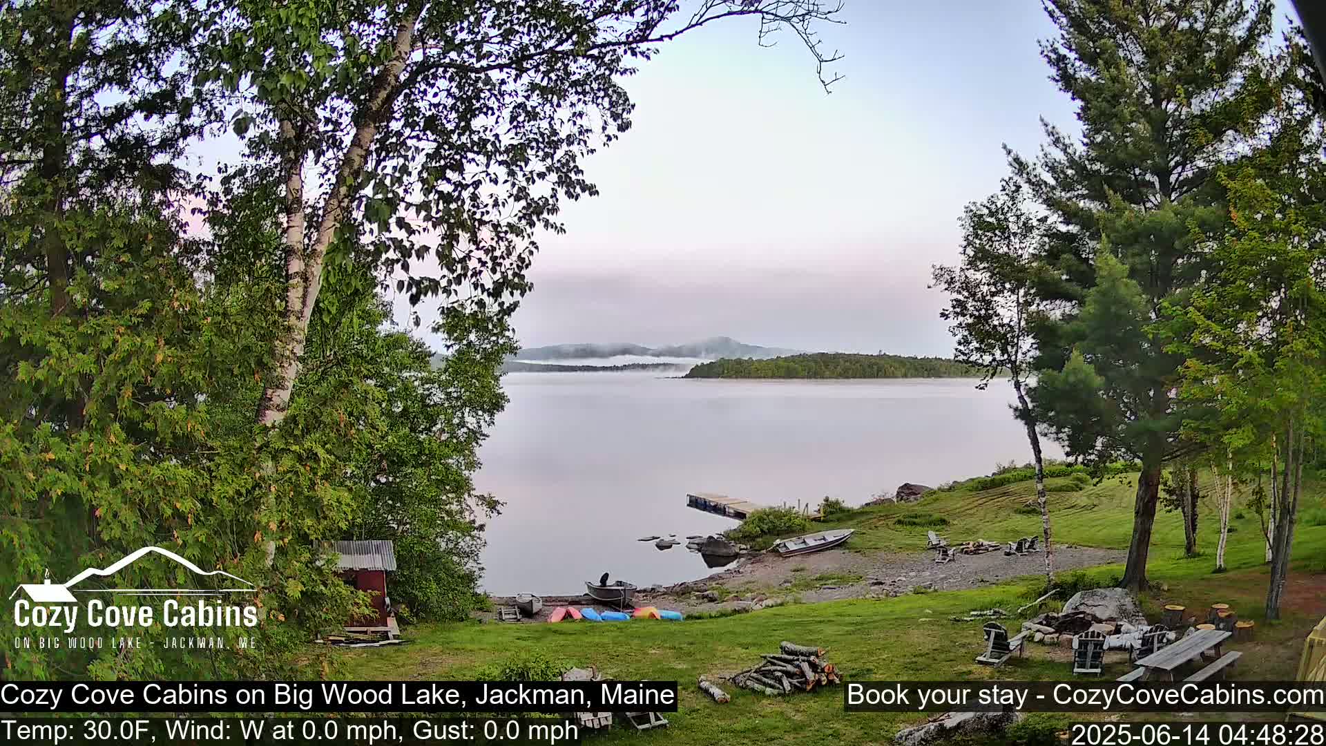 A calm lake with a misty shoreline is seen from a lakeside property with several kayaks and a small boat on the shore, under a partly cloudy sky.