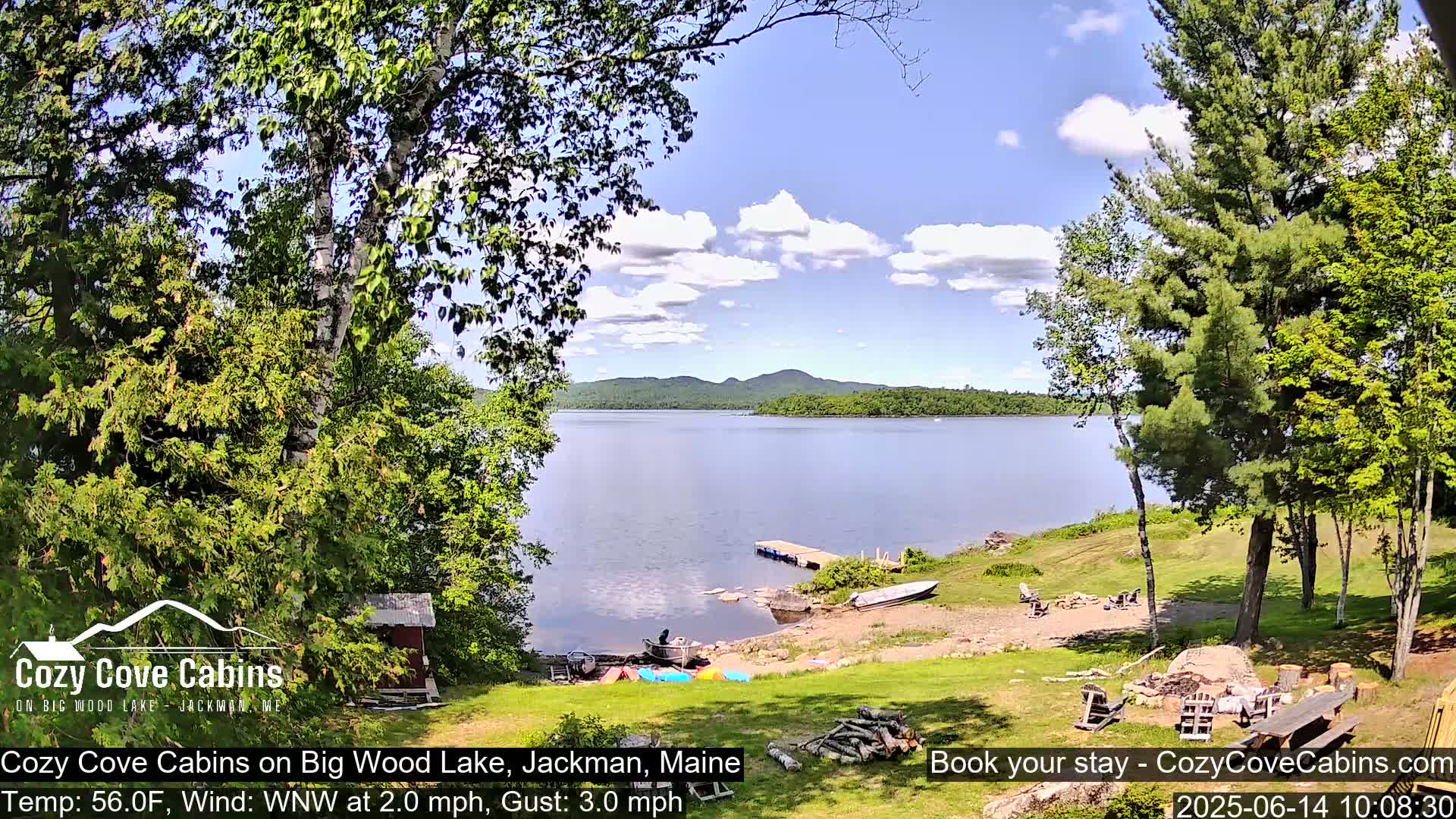 A lakeside view shows a calm lake under a partly cloudy sky, with several boats on the shore near a small cabin and trees.
