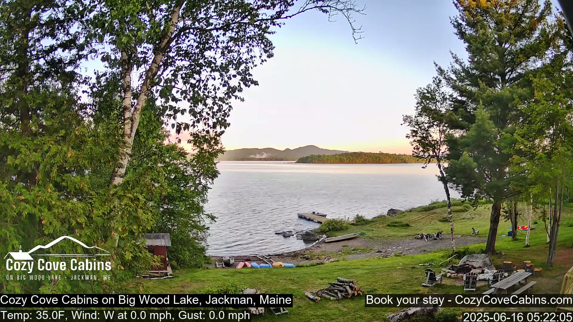 A calm lake at sunset is viewed from a grassy lakeside yard with trees, a small dock, and canoes.