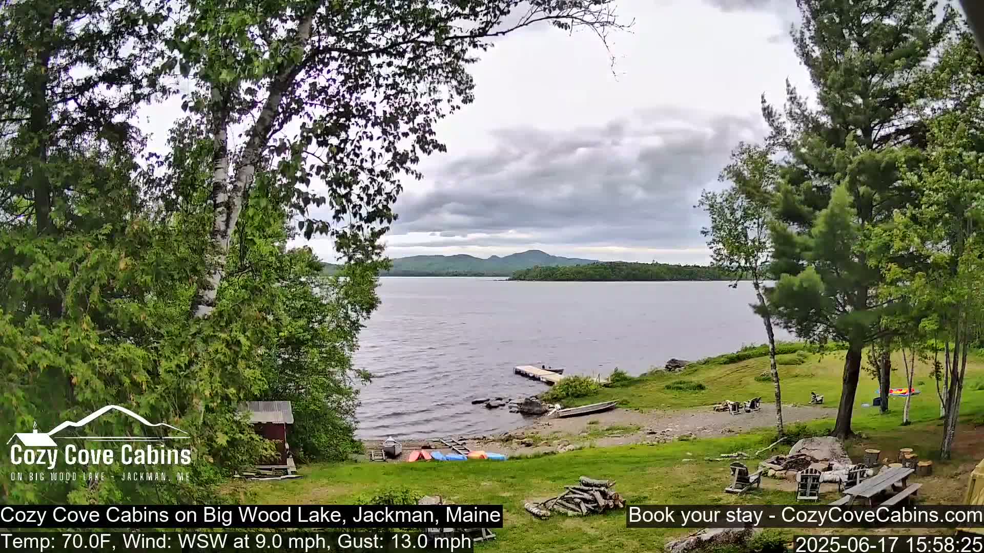 A lakeside view shows a calm lake under a cloudy sky, with trees, a small dock, and some recreational equipment near a sandy shoreline.