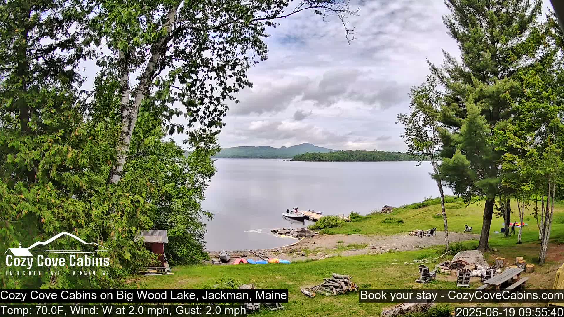 A tranquil lakeside scene with a partly cloudy sky, featuring a calm lake, trees, a small dock with boats, and a grassy area with outdoor furniture.