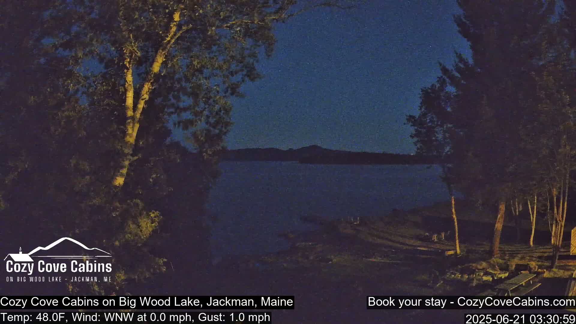 A nighttime view of a calm lake under a dark sky, with trees and shoreline visible in the foreground.
