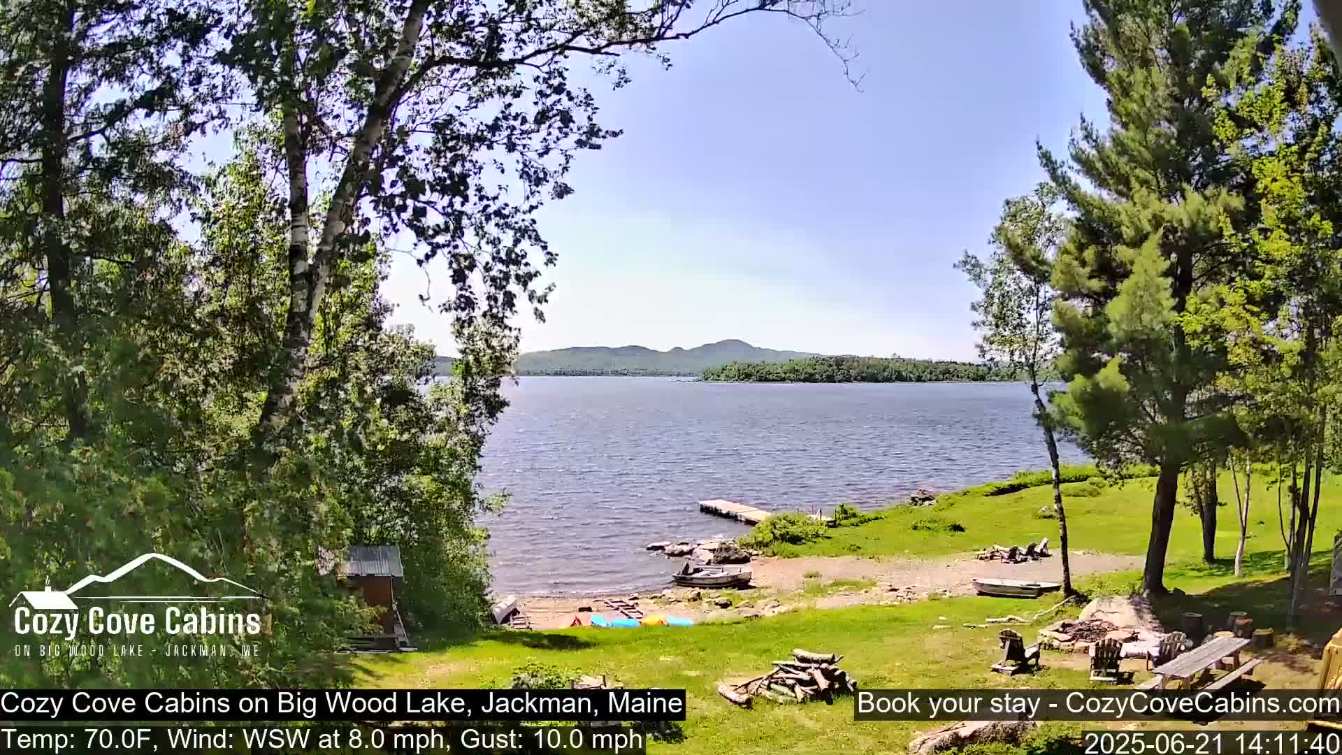 A calm lake with a sandy shore and several boats, surrounded by lush green trees under a clear blue sky, with a gentle breeze.