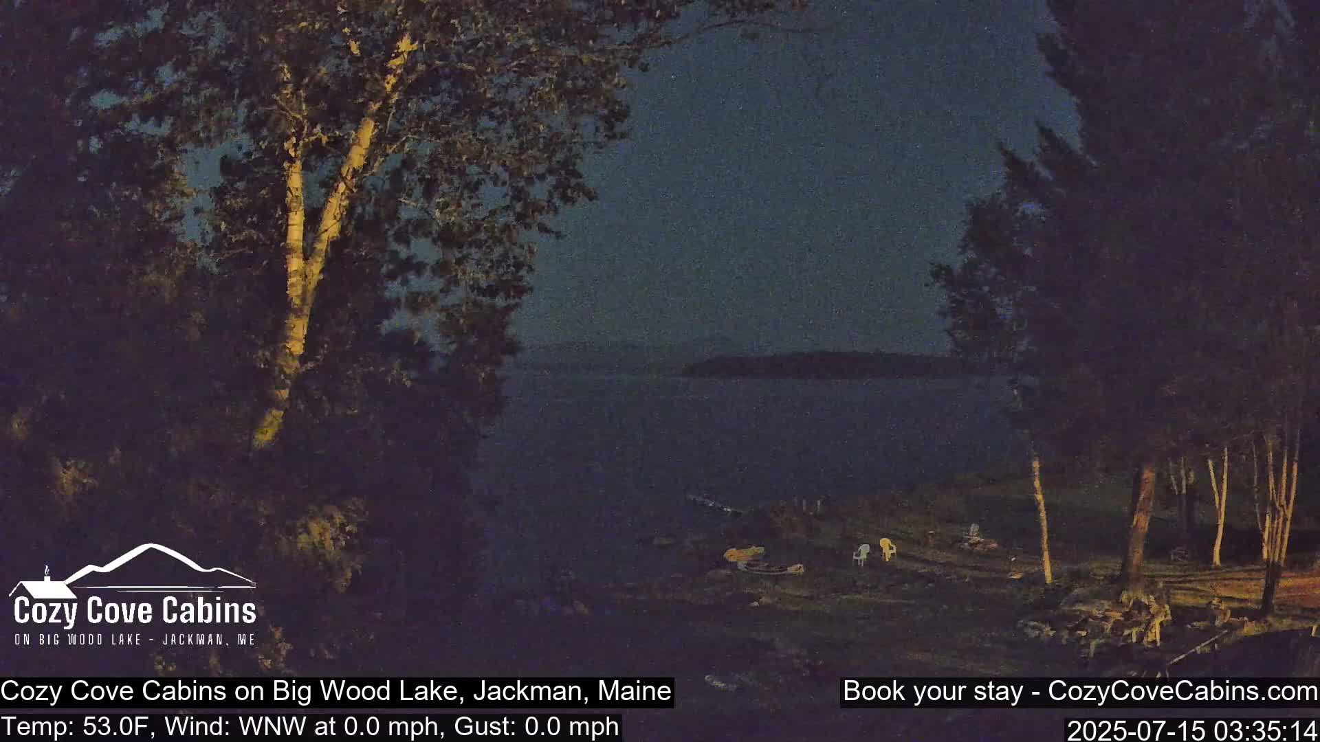 A dark, calm night scene shows a lake with an island in the distance, trees lining the shore, and a few chairs near the water's edge.