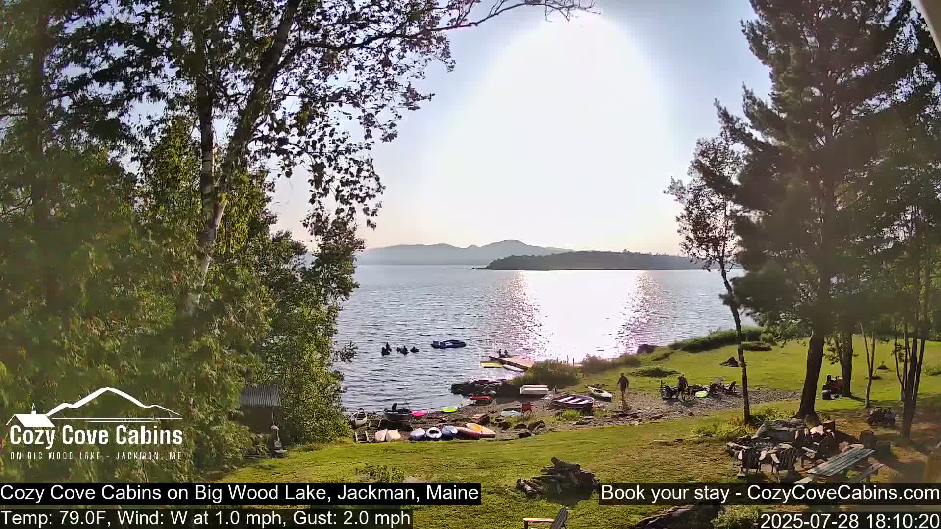 A sunny lakeside scene shows calm water with several small boats and people gathered on a grassy shore near trees.