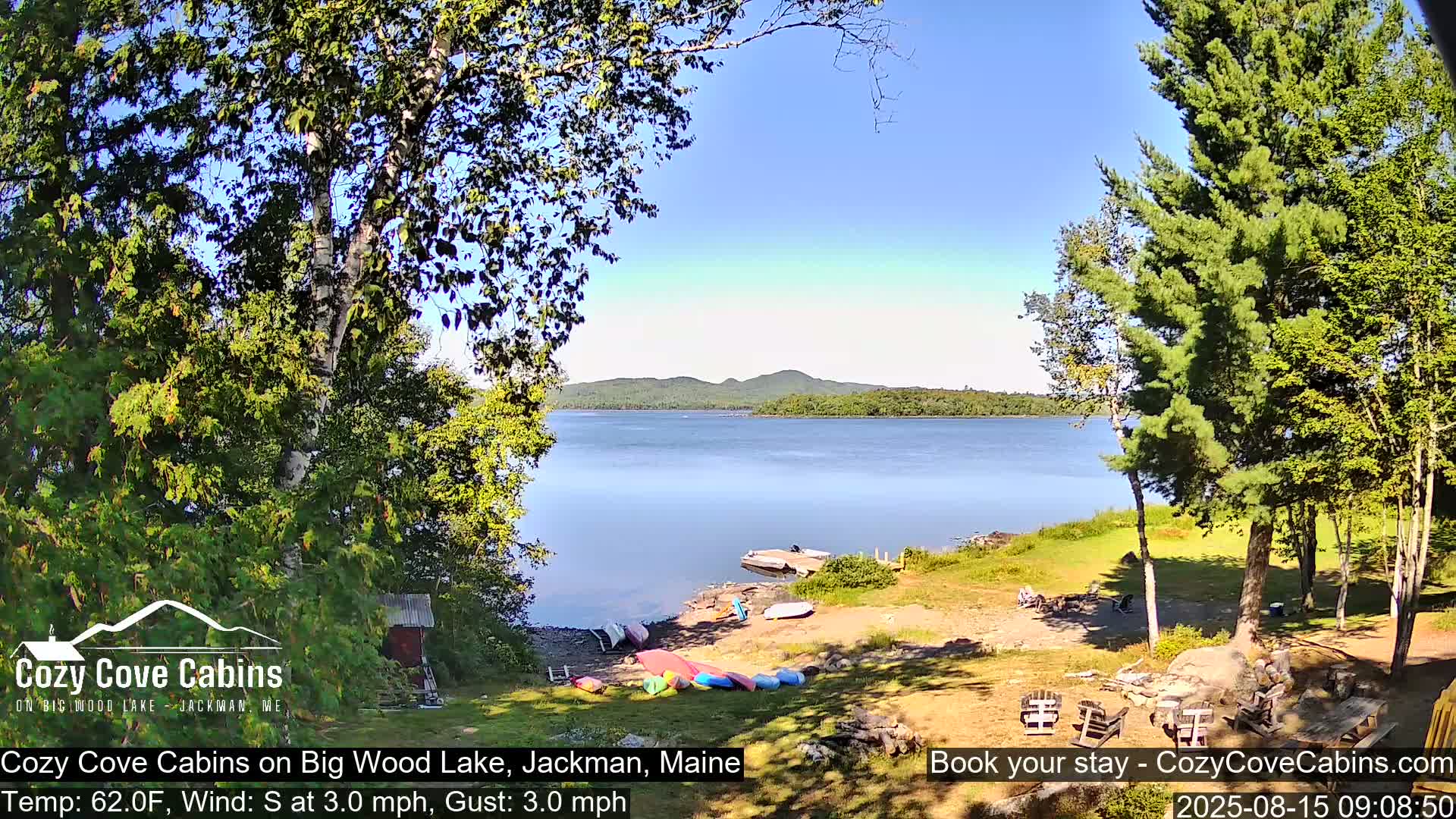 A sunny lakeside scene shows calm water with several small boats and people gathered on a grassy shore near trees.