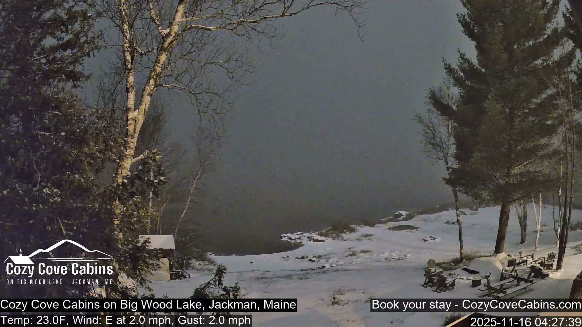 A cold, hazy winter scene at night or early morning shows a snow-covered shoreline with evergreens and bare trees framing a dark, still lake, with outdoor seating dusted in snow.