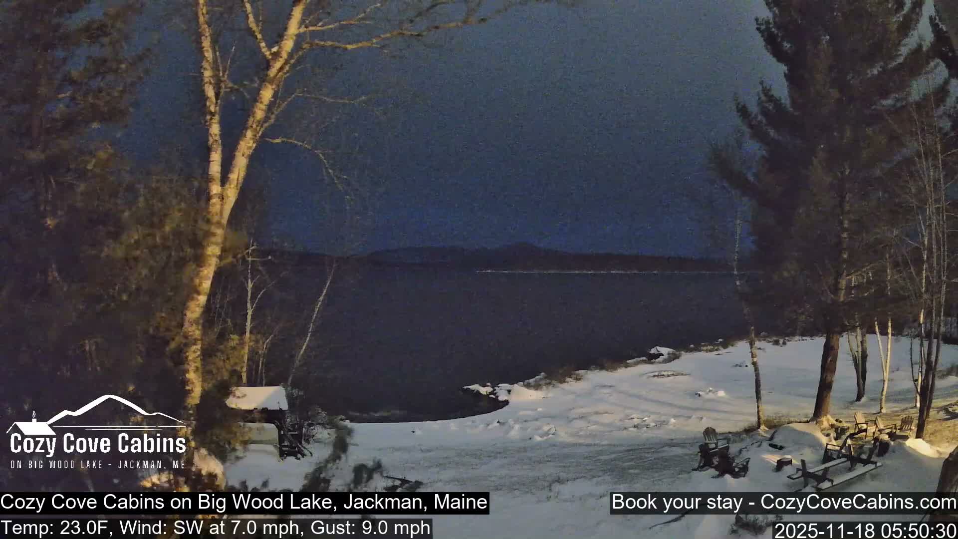 A cold, hazy winter scene at night or early morning shows a snow-covered shoreline with evergreens and bare trees framing a dark, still lake, with outdoor seating dusted in snow.
