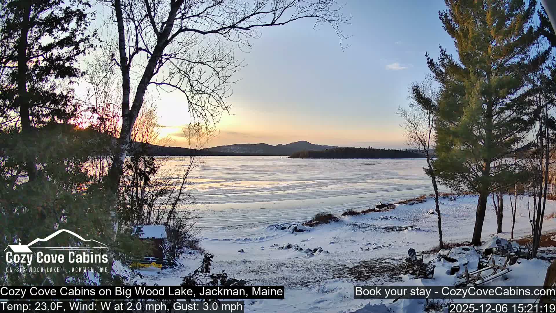 A clear, cold winter landscape features a vast, mostly frozen lake reflecting a colorful twilight sky, bordered by snow-covered ground with evergreen and deciduous trees and distant mountains.