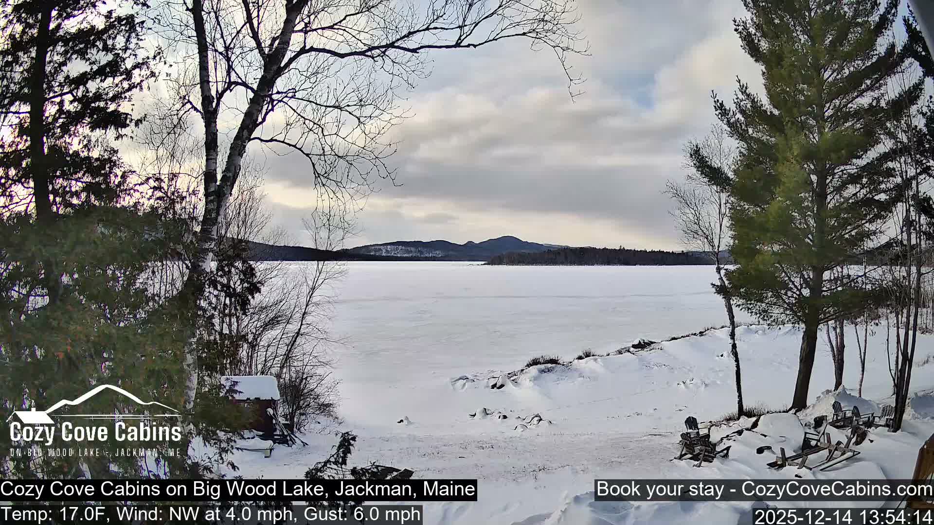 A clear, cold winter landscape features a vast, mostly frozen lake reflecting a colorful twilight sky, bordered by snow-covered ground with evergreen and deciduous trees and distant mountains.