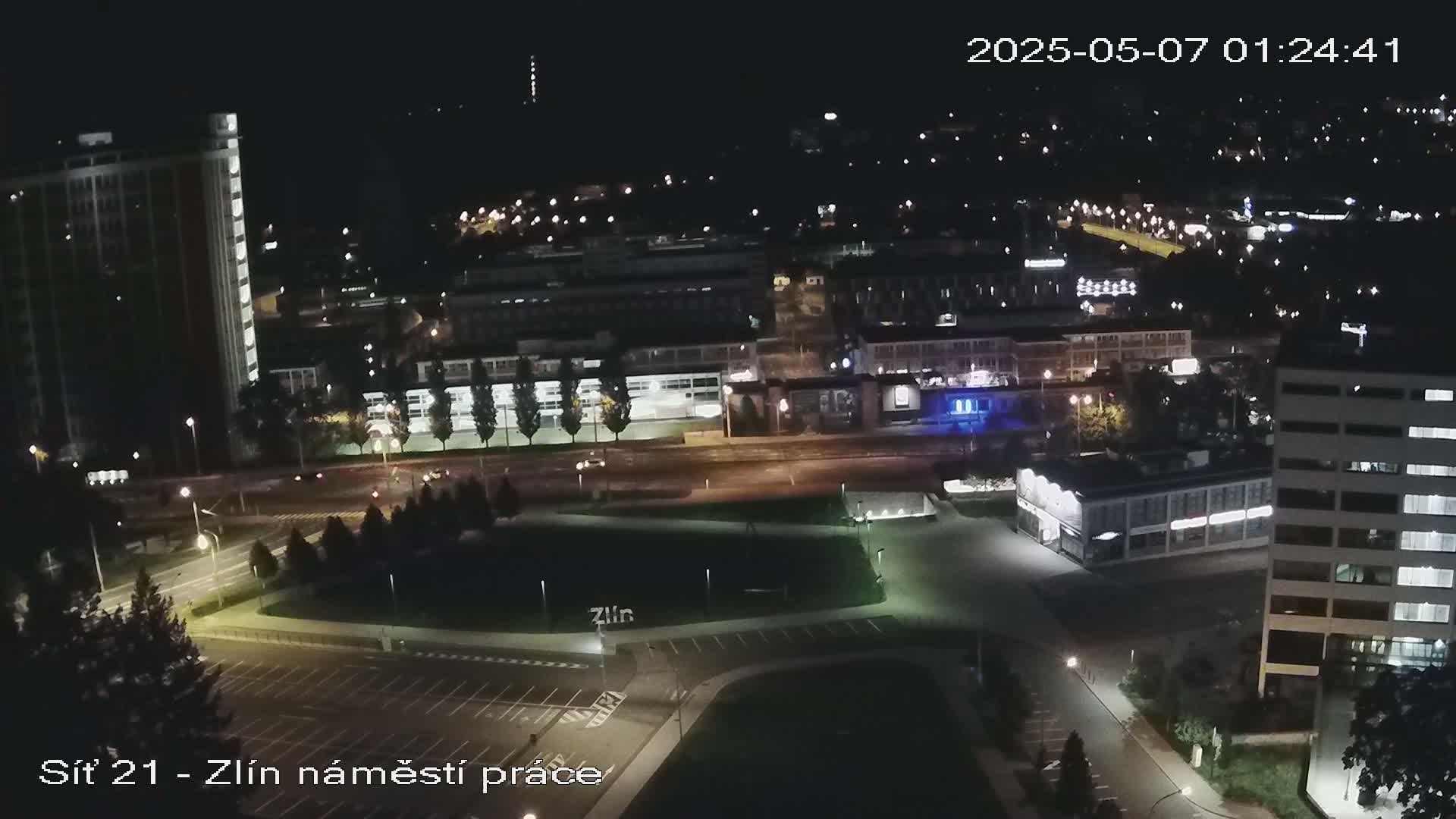 A nighttime aerial view of a city shows roads, buildings, and a park, illuminated by artificial lights.