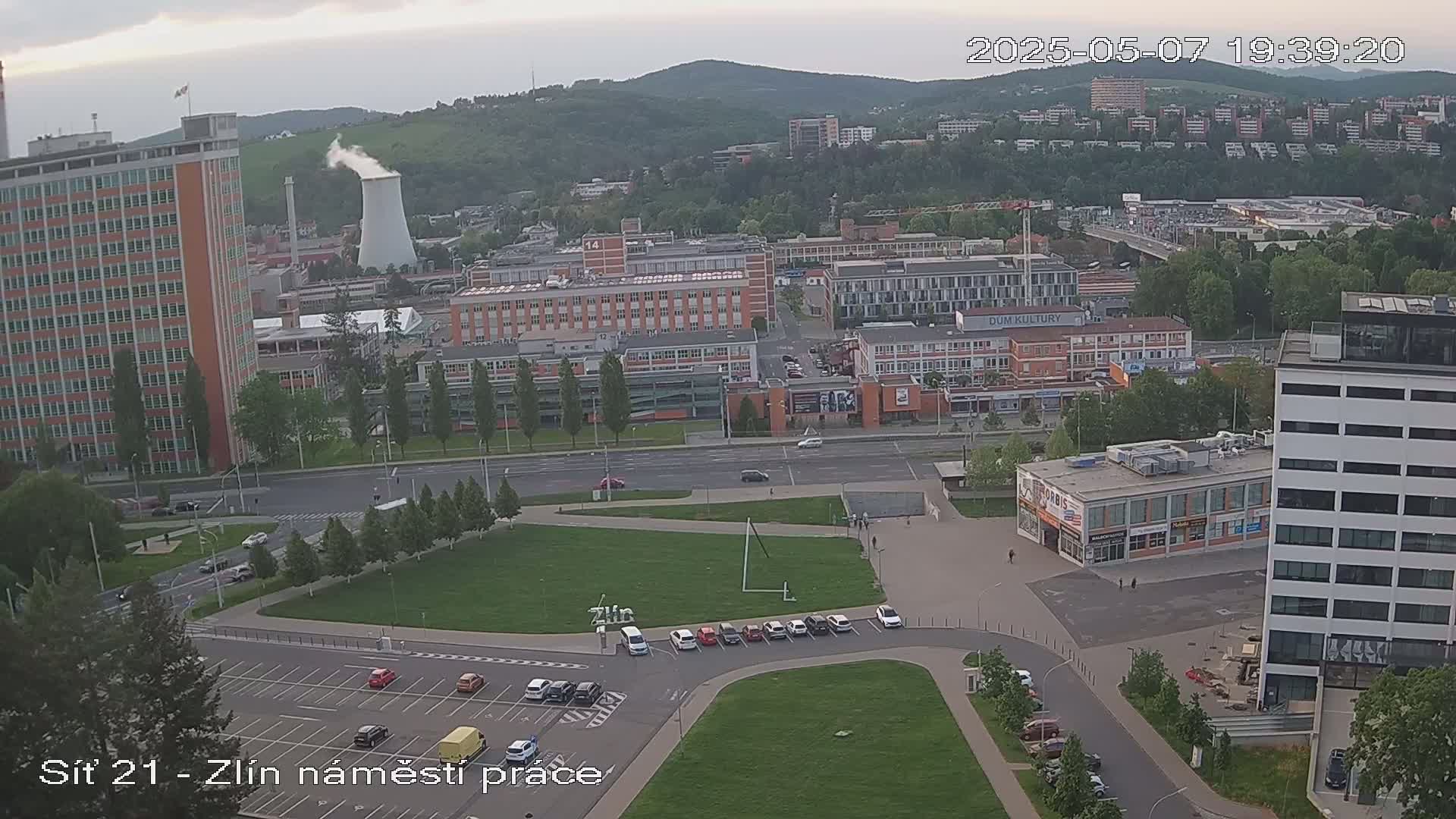 An overcast day reveals a city scene with numerous buildings, including tall apartment buildings, industrial structures, and a large parking lot, all situated in a valley against a backdrop of hills.