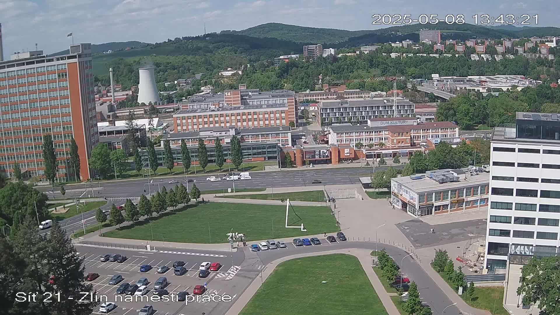 A high-angle view of a city on a partly cloudy day, showing a large grassy area with buildings and parking lots surrounding it.