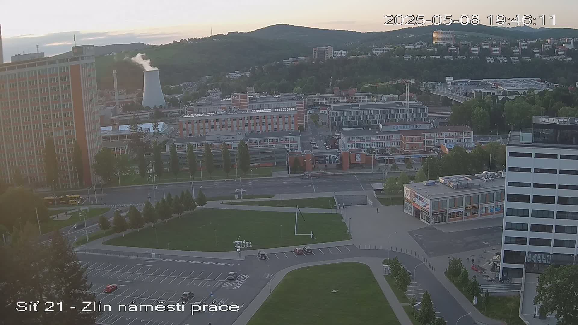 An aerial view shows a city scene at dusk, featuring buildings, roads, a grassy area, and a tall industrial smokestack emitting smoke.