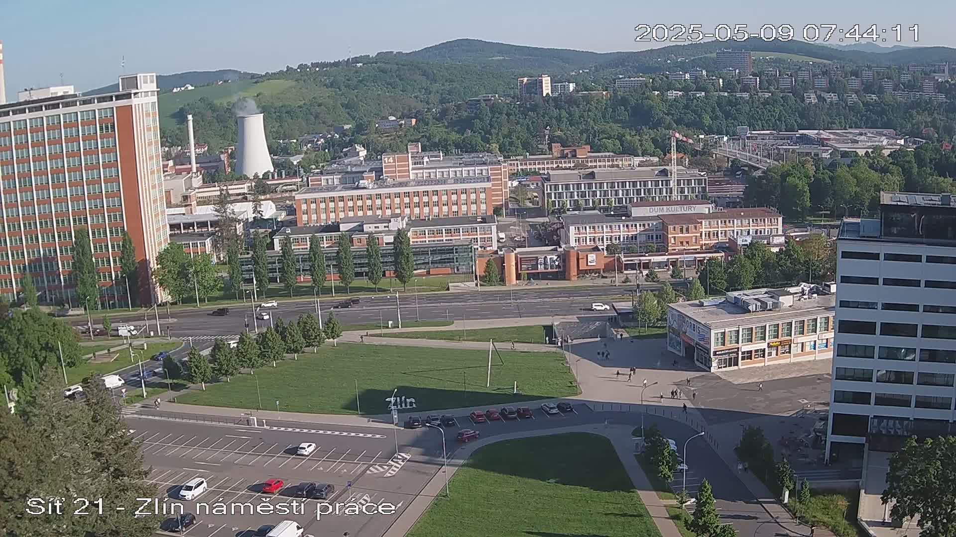 A high-angle, long shot on a partly sunny day shows a city scene with many buildings, a large grassy area, and a parking lot, all surrounded by trees and hills in the background.