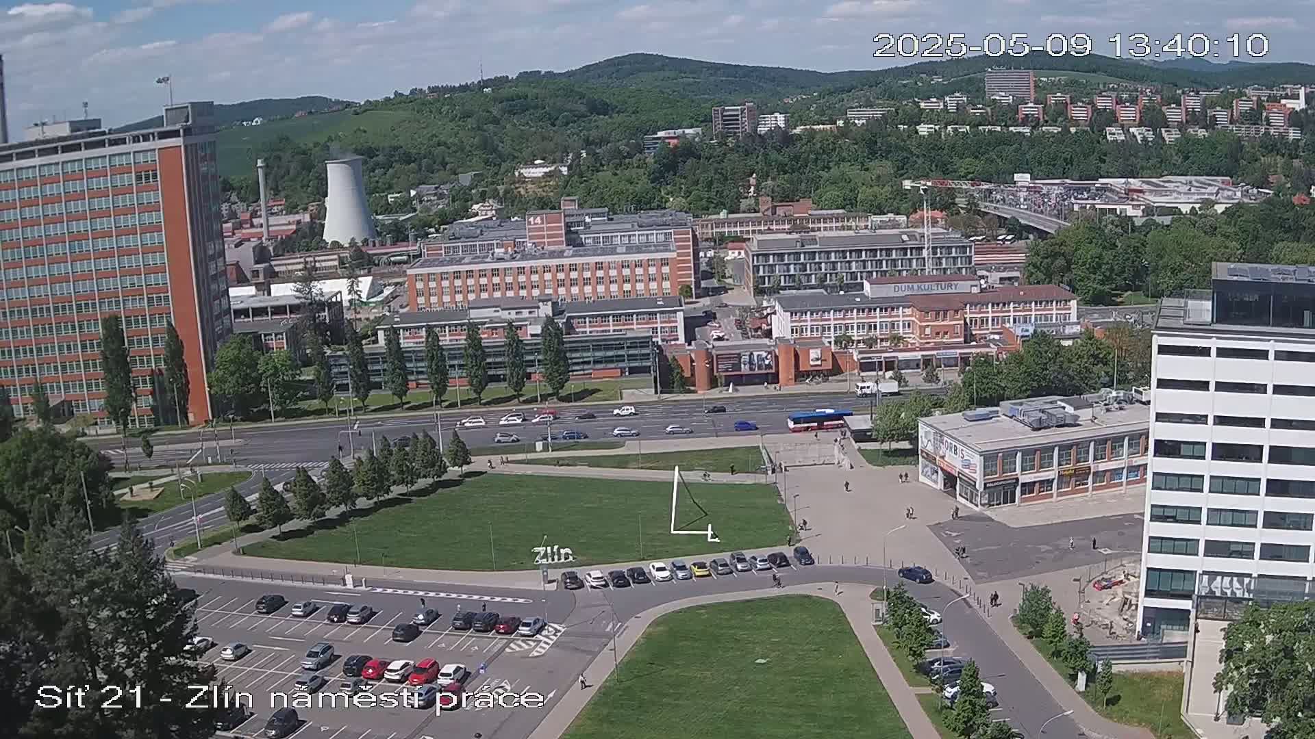 A high-angle view of a city scene on a partly cloudy day, showing several buildings, a grassy plaza with parked cars, and a road with vehicles.