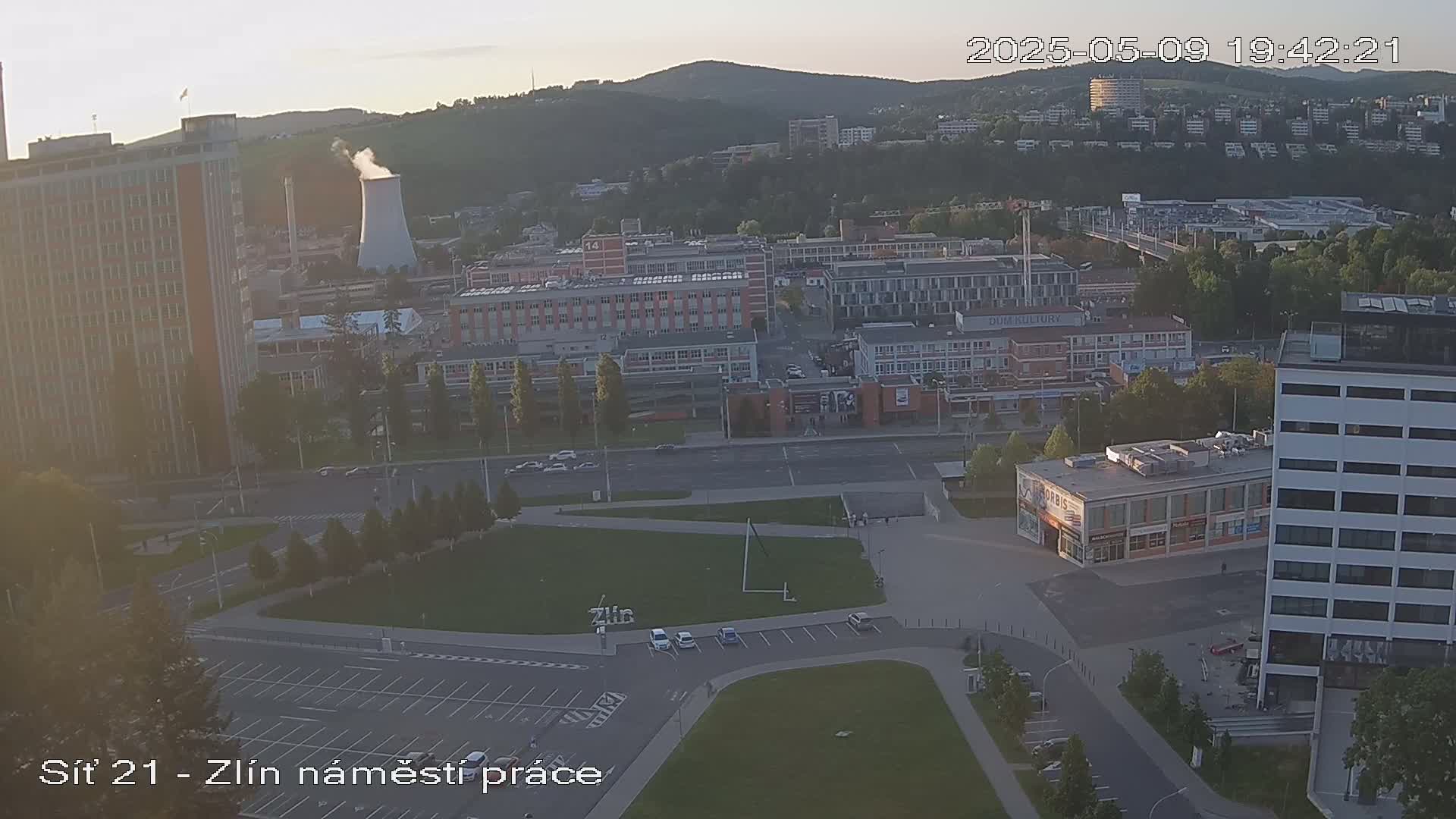 An aerial view shows a city scene at dusk, with several buildings, a parking lot, and a grassy area, under a mostly clear sky.