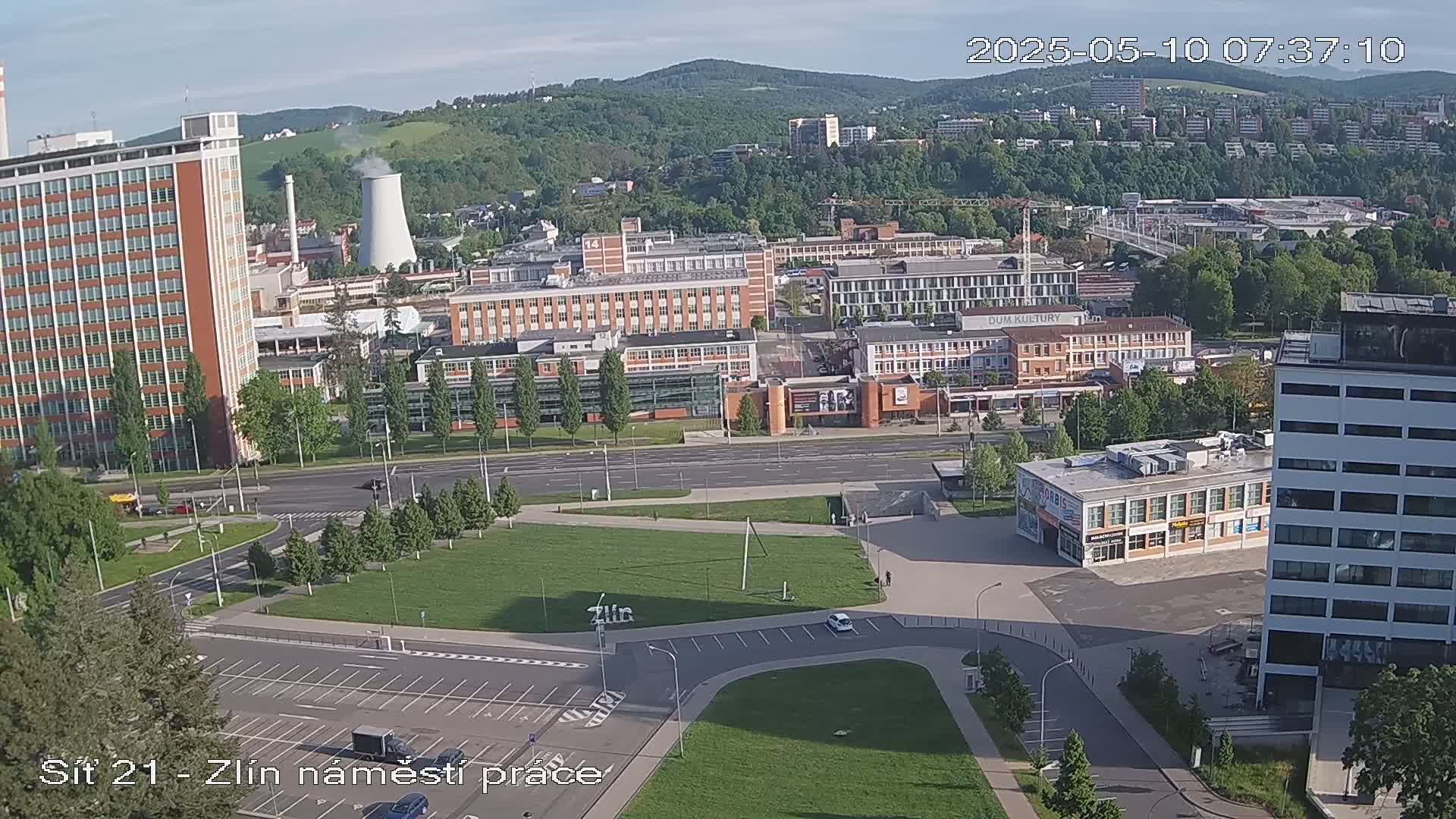 A high-angle, wide shot of a city on a partly sunny day, showing a large square with parking lots, various buildings including high-rises and industrial structures, and green spaces.