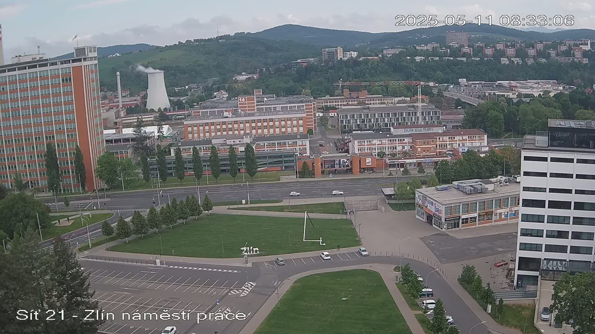 An aerial view of a city scene on an overcast day, showing several buildings, a large grassy area, and a parking lot, with a few cars scattered throughout.