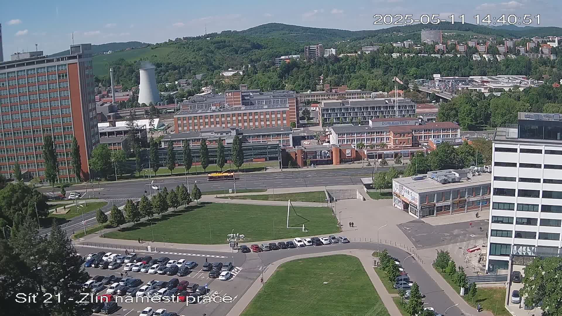 An aerial view of a city block on a sunny day shows several buildings, parking lots full of cars, a grassy area, and a road with some traffic.