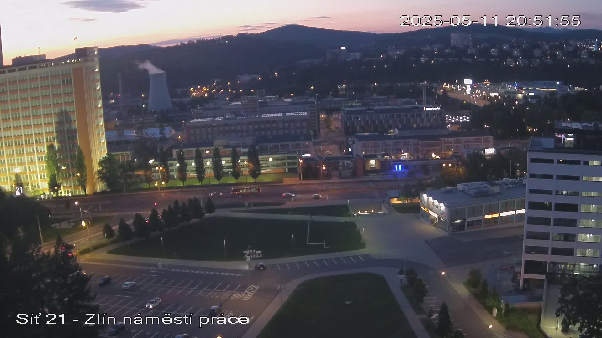 An elevated, twilight view of a city with several buildings, streets, and a large grassy area, under a mostly clear sky.