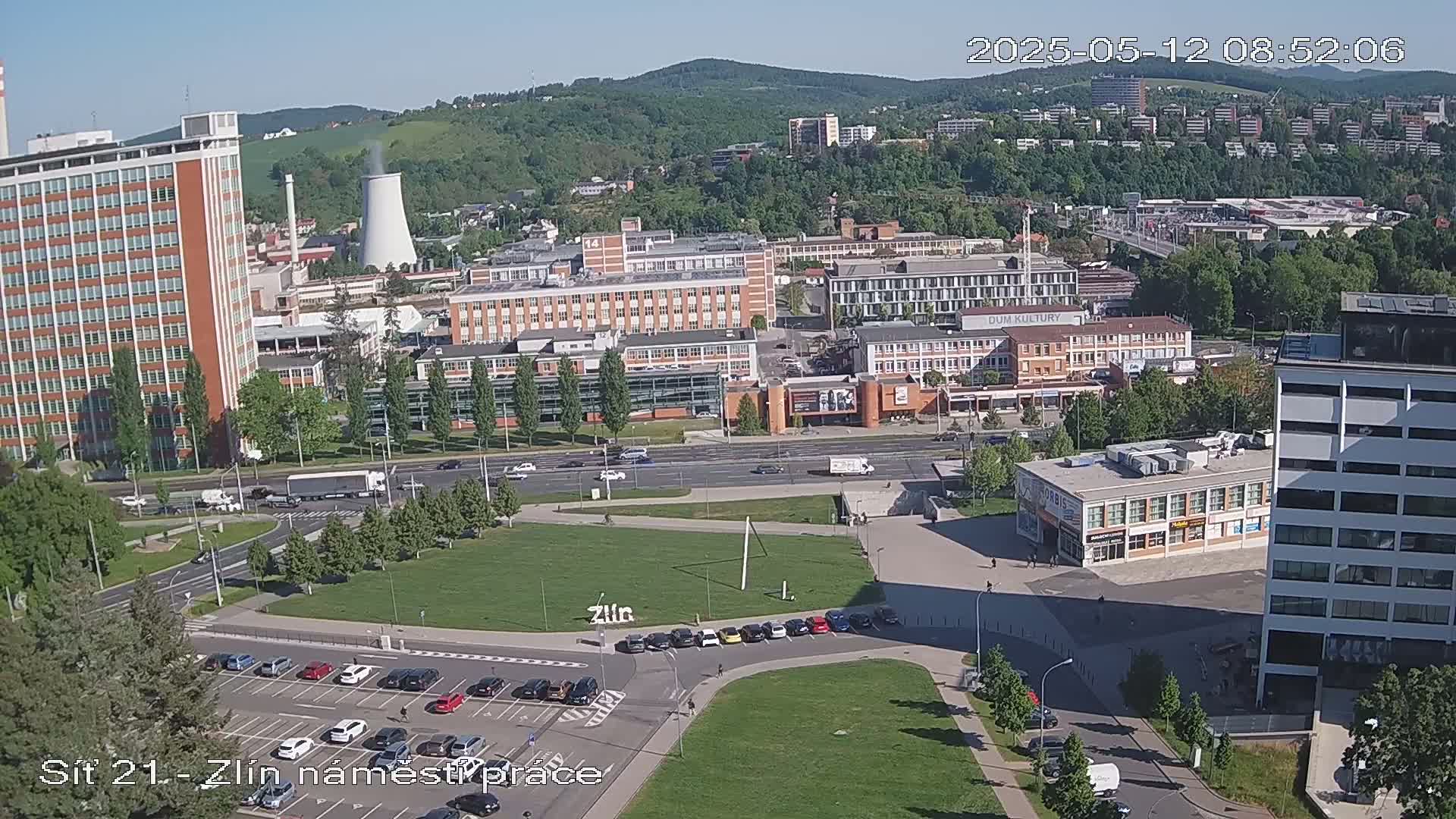 An aerial view of a city on a sunny day, showing several large buildings, a grassy park, and a busy roadway.