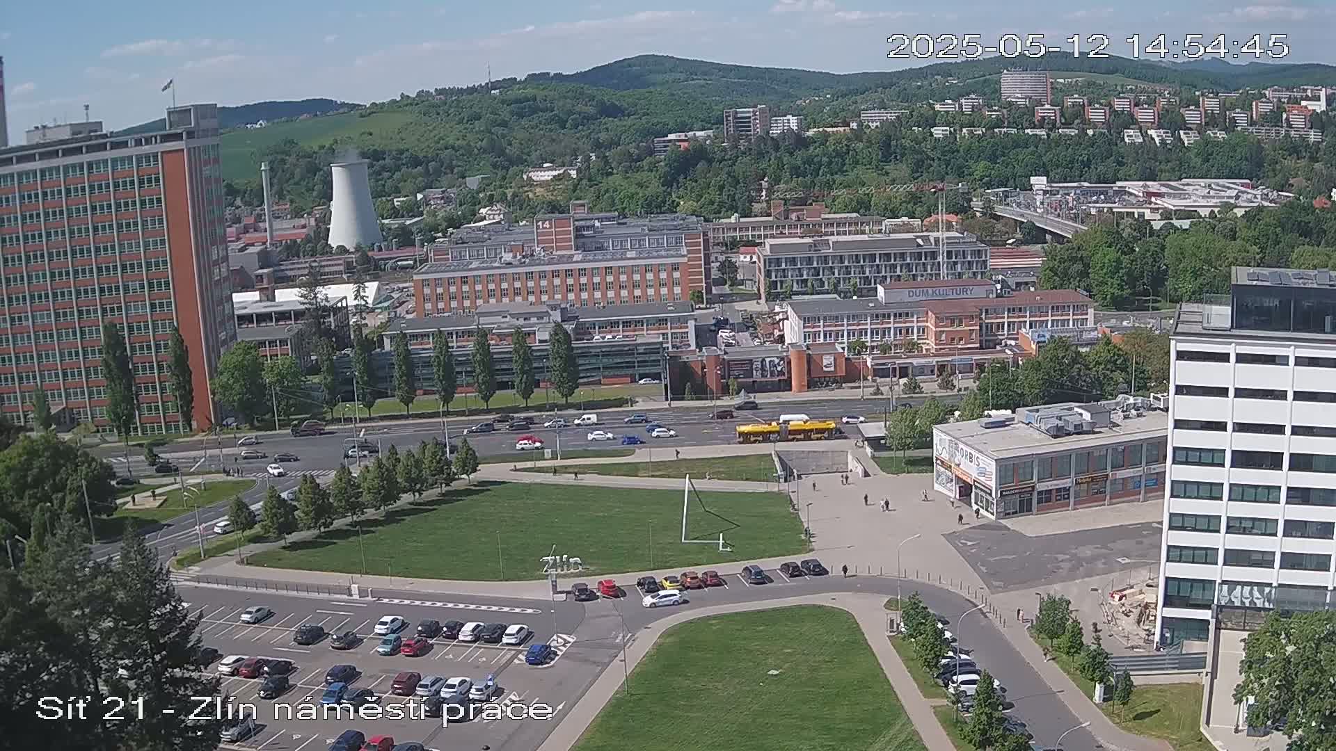 A sunny aerial view of a city with numerous buildings, a grassy park, a roadway with cars, and a parking lot.