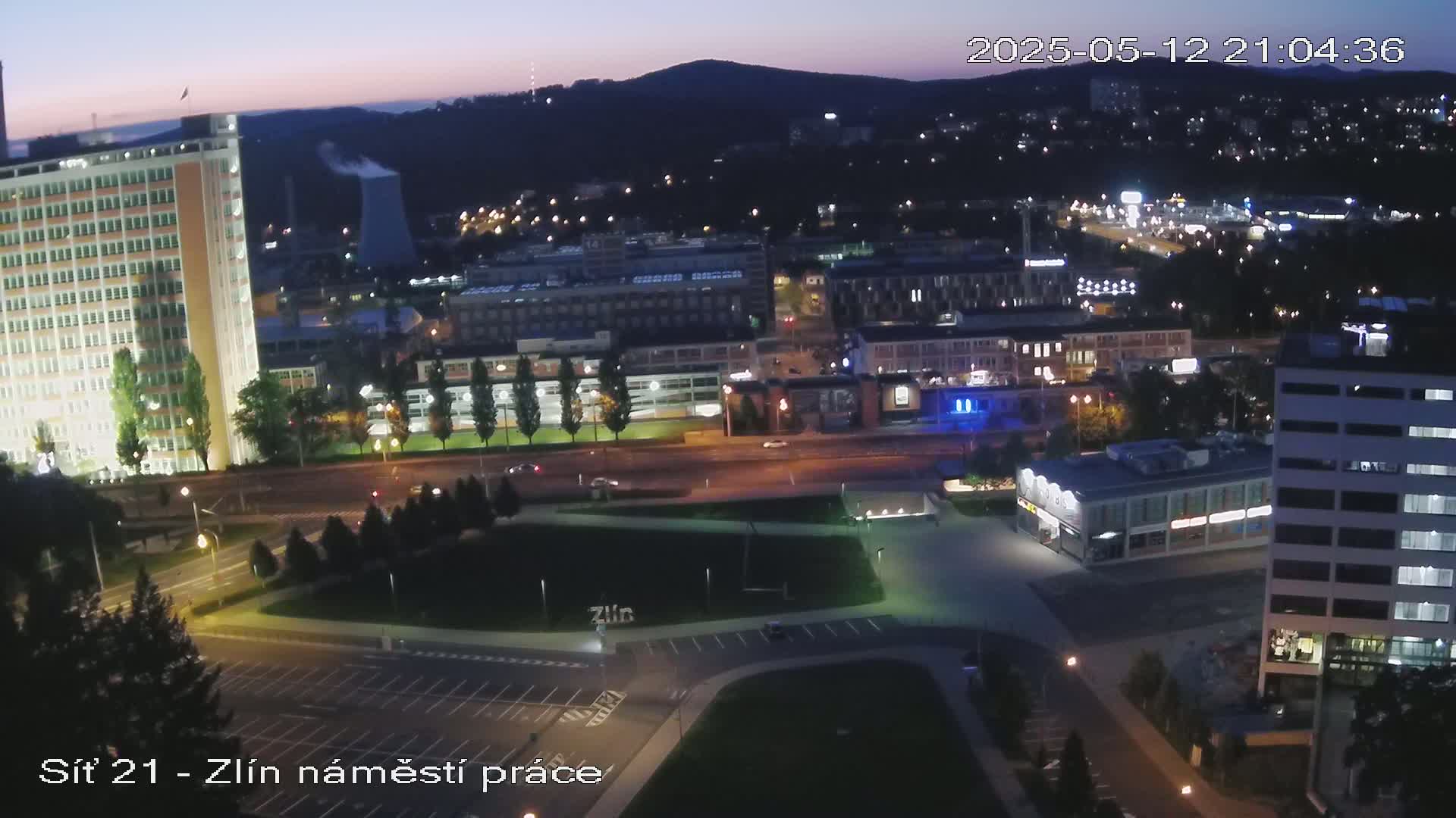 A nighttime, high-angle view of a city, showing illuminated buildings, roads, and a park-like area with some trees.