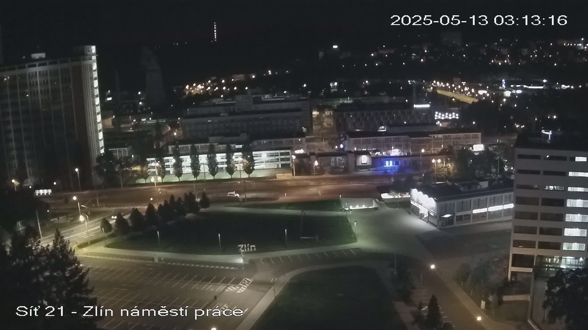 A nighttime aerial view of a city with several buildings and a large empty square, illuminated by streetlights.