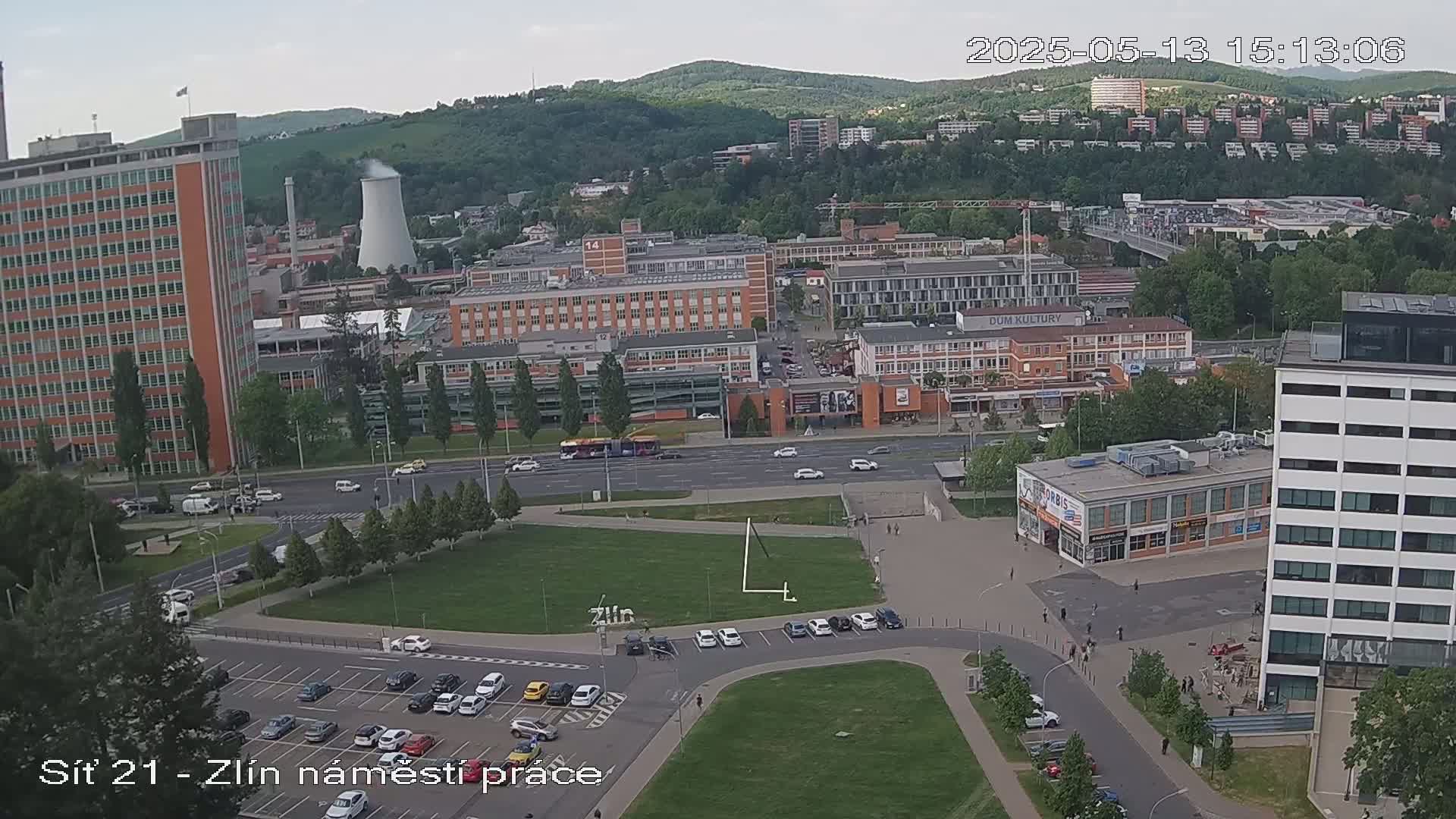 A high-angle, long shot of a city scene on a partly cloudy day, showing several buildings, streets with cars, a grassy area with parked cars, and a large industrial structure in the distance.