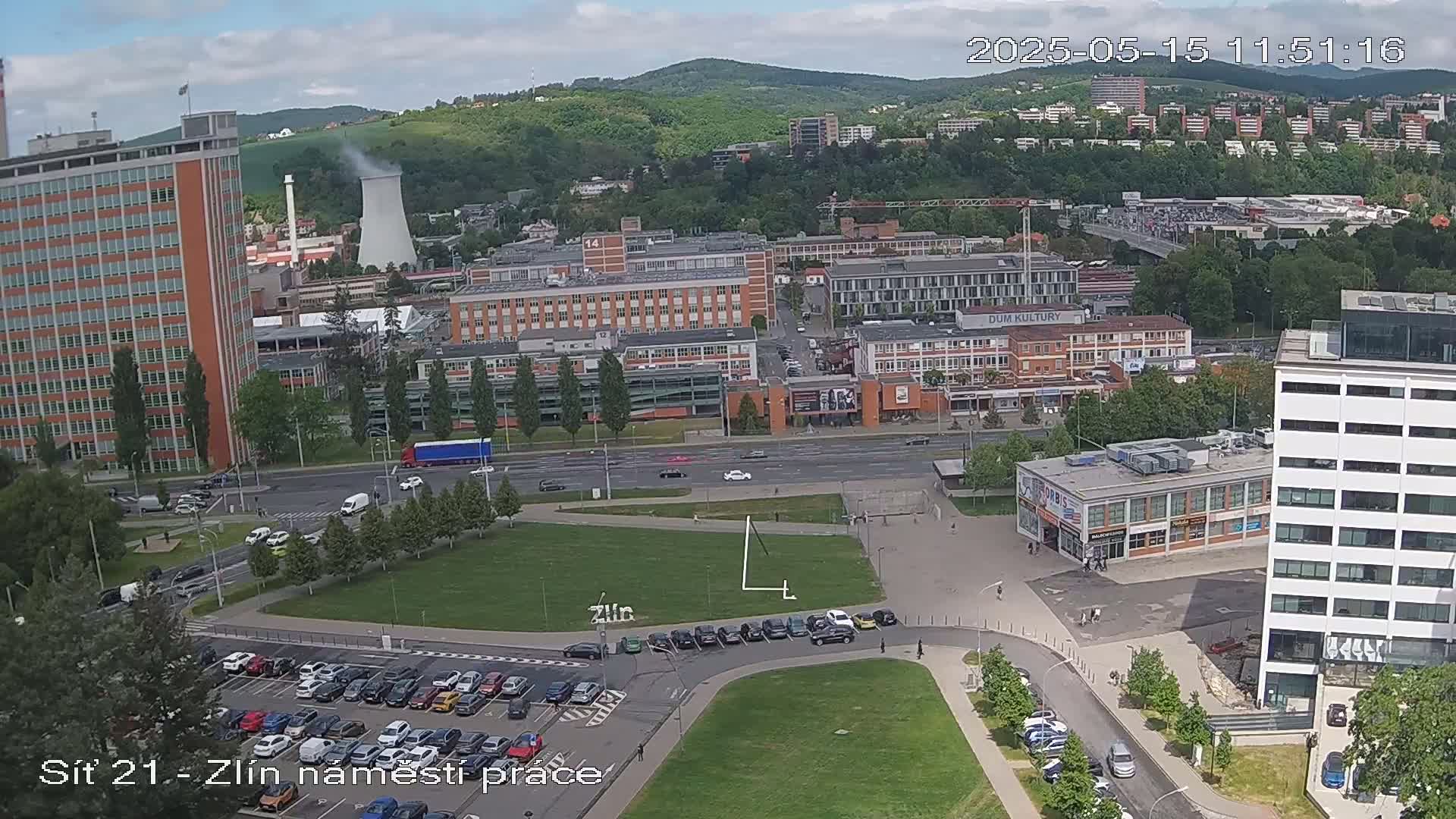 An aerial view shows a city scene on a partly cloudy day, with numerous buildings, vehicles, and a large grassy area, featuring a parking lot and a road.