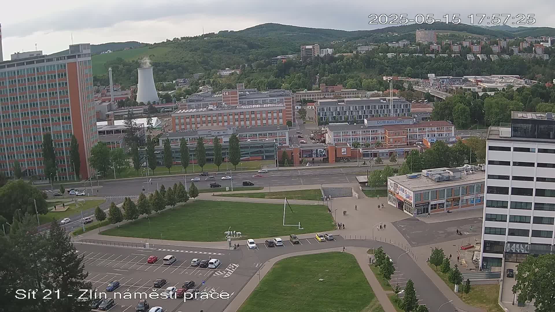 An aerial view shows a city under partly cloudy skies, featuring several buildings, a large grassy area with parked cars, and a cooling tower in the distance.