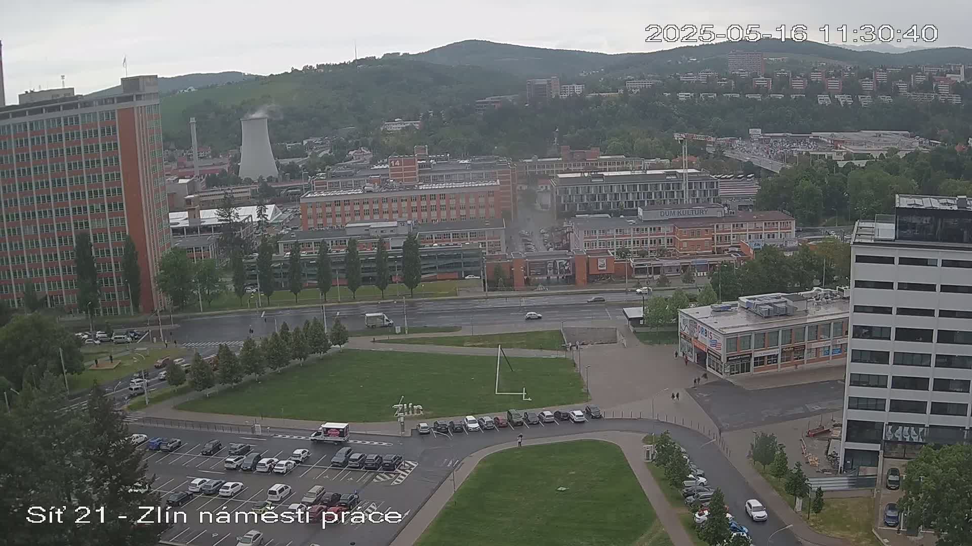 An overcast day reveals a city scene with several buildings, a large grassy area, parking lots, and roads, all surrounded by greenery and distant hills.