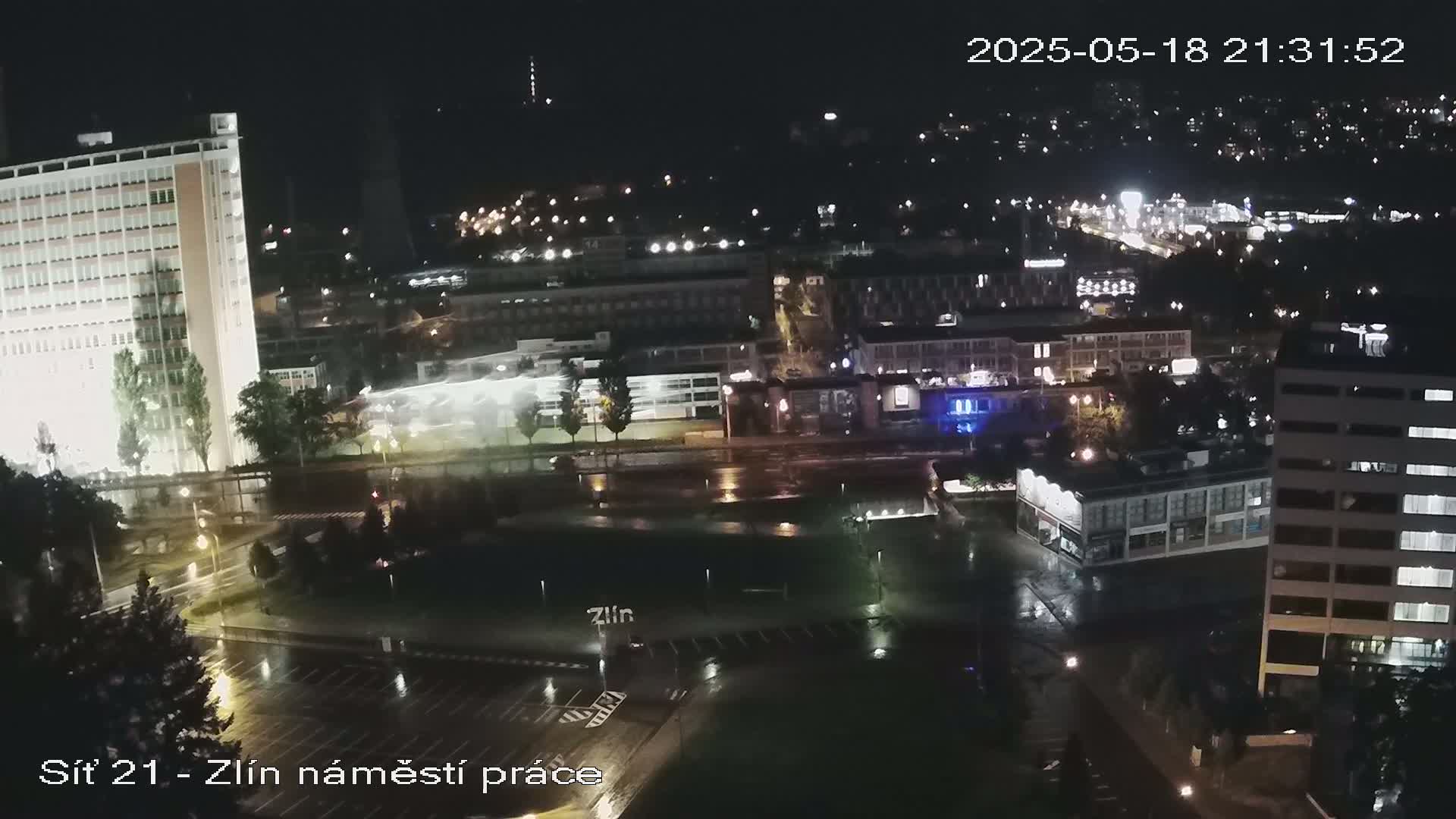 A nighttime view of a city with wet streets and illuminated buildings under rainy conditions.