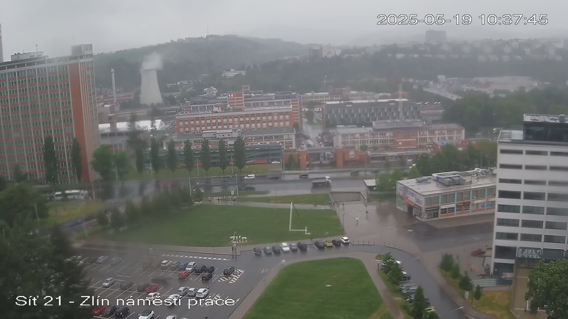 A high-angle view of a city landscape during a rainy day, showing several buildings, a parking lot, and a large grassy area.