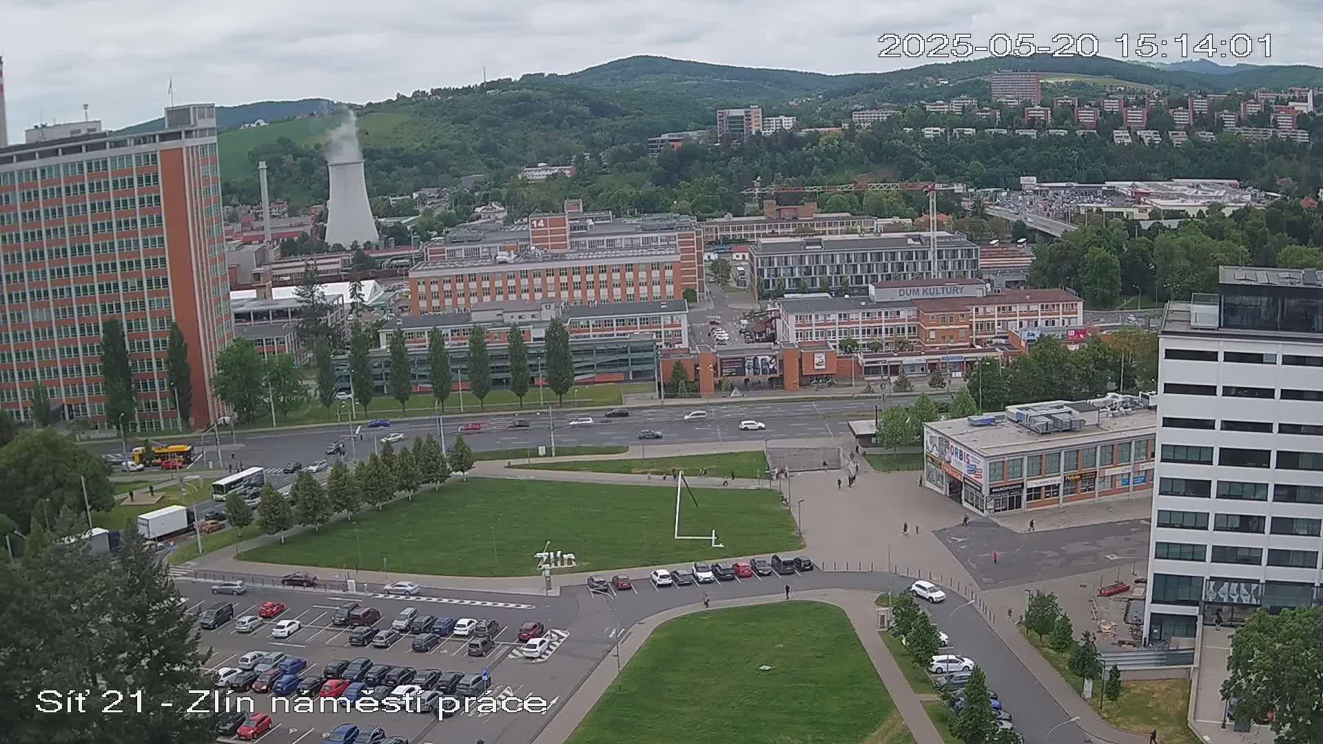 An aerial view shows a city square with a parking lot, several multi-story buildings, and a large grassy area under an overcast sky.