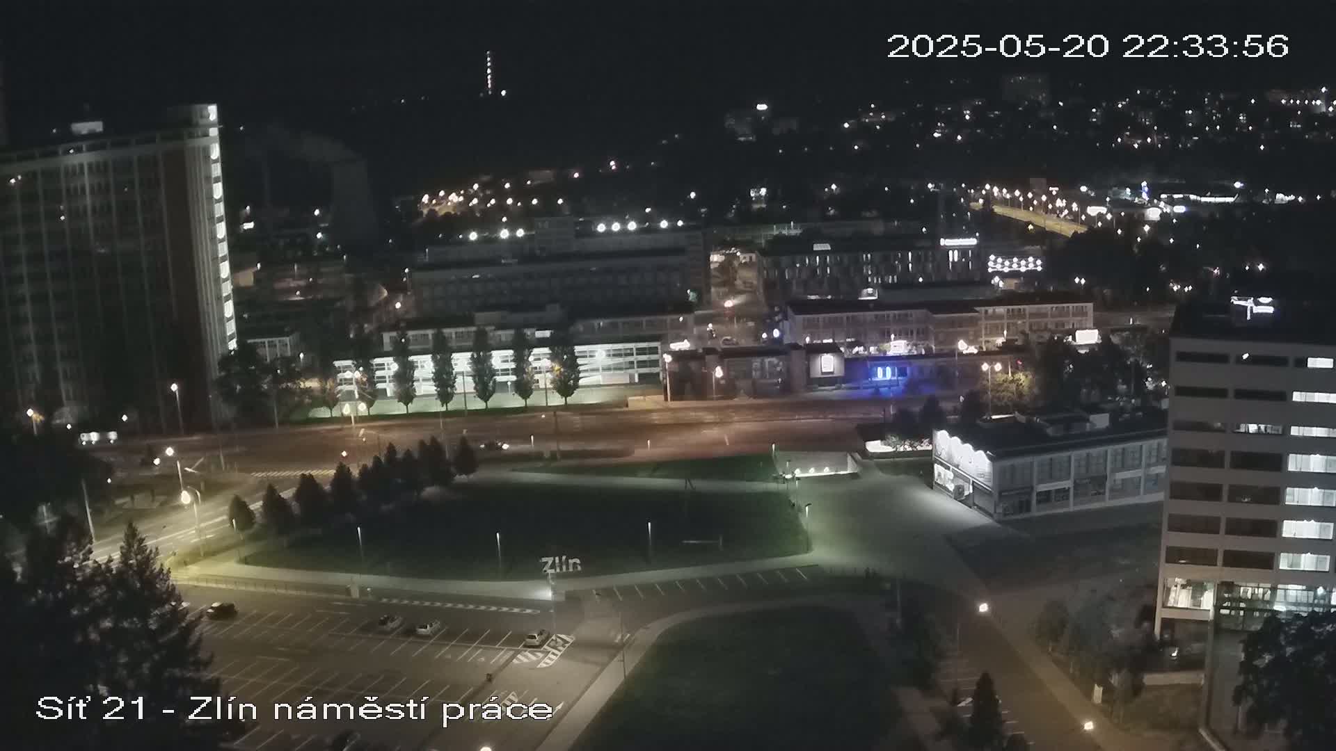 A nighttime aerial view of a city showing buildings, roads, and a park with cars parked in a lot.