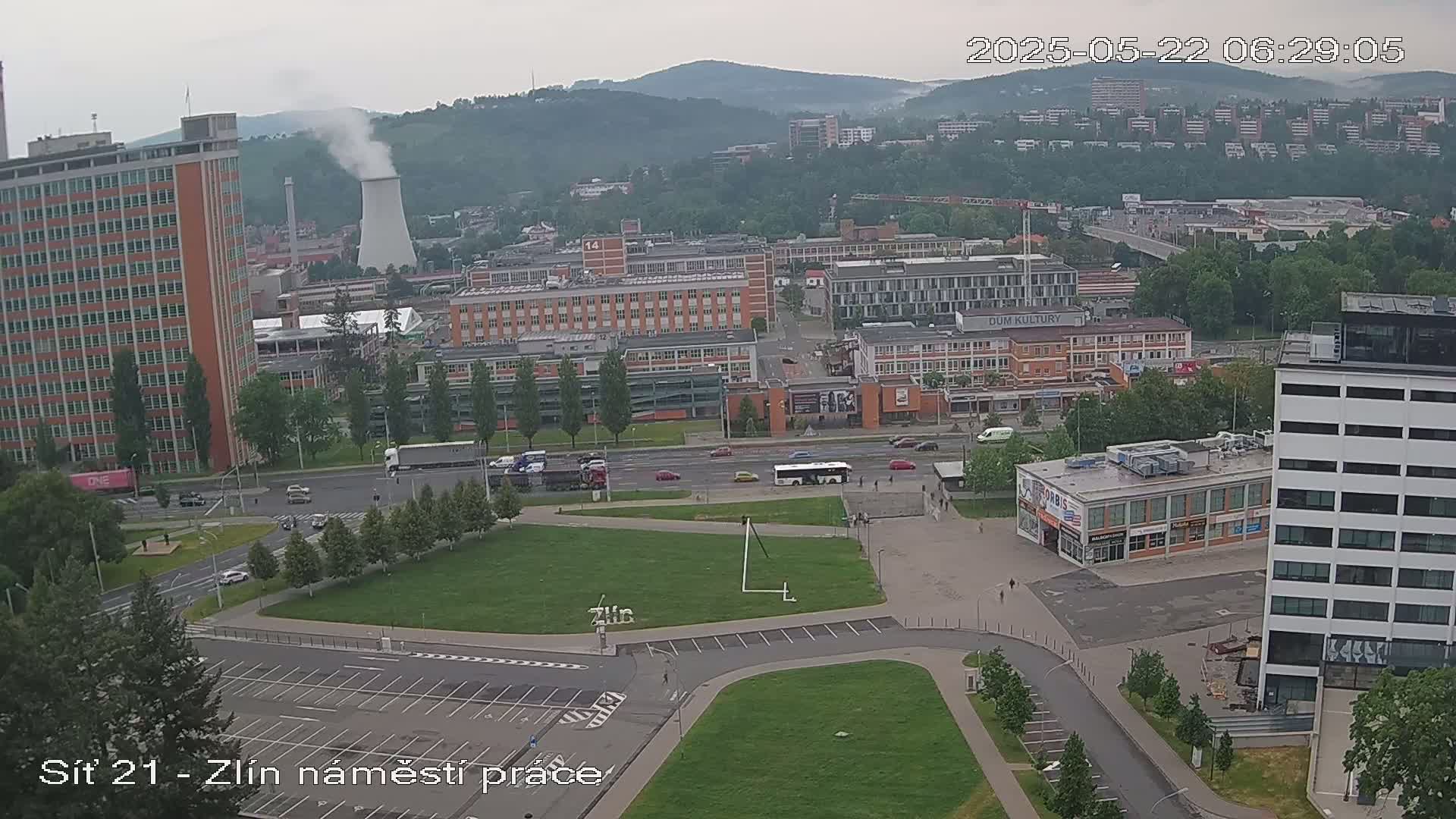 A city scene on an overcast day shows a large grassy area with a parking lot, roads with cars, numerous buildings, and a smokestack emitting smoke in the background.