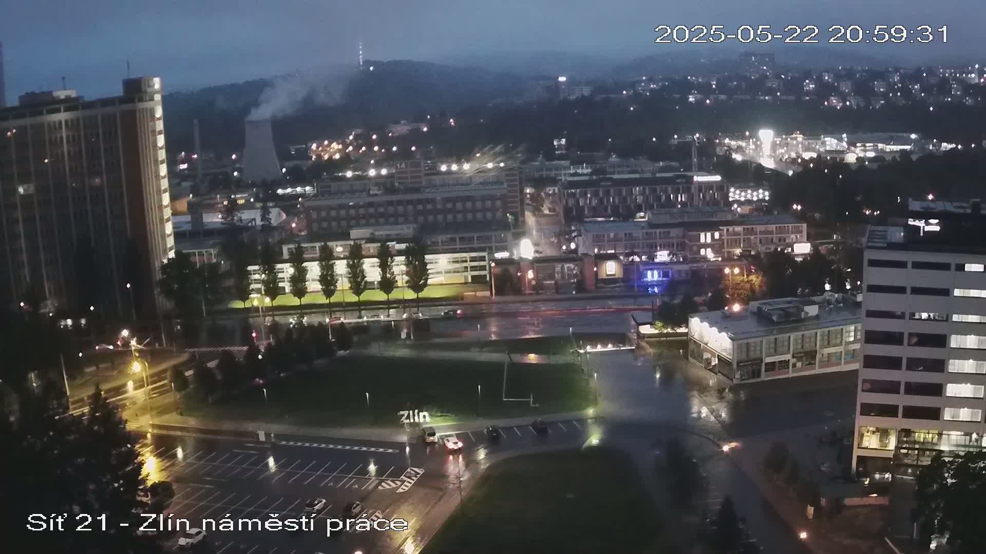 A nighttime aerial view of a city during a rain shower, showing illuminated streets, buildings, and a large open area with some vehicles.