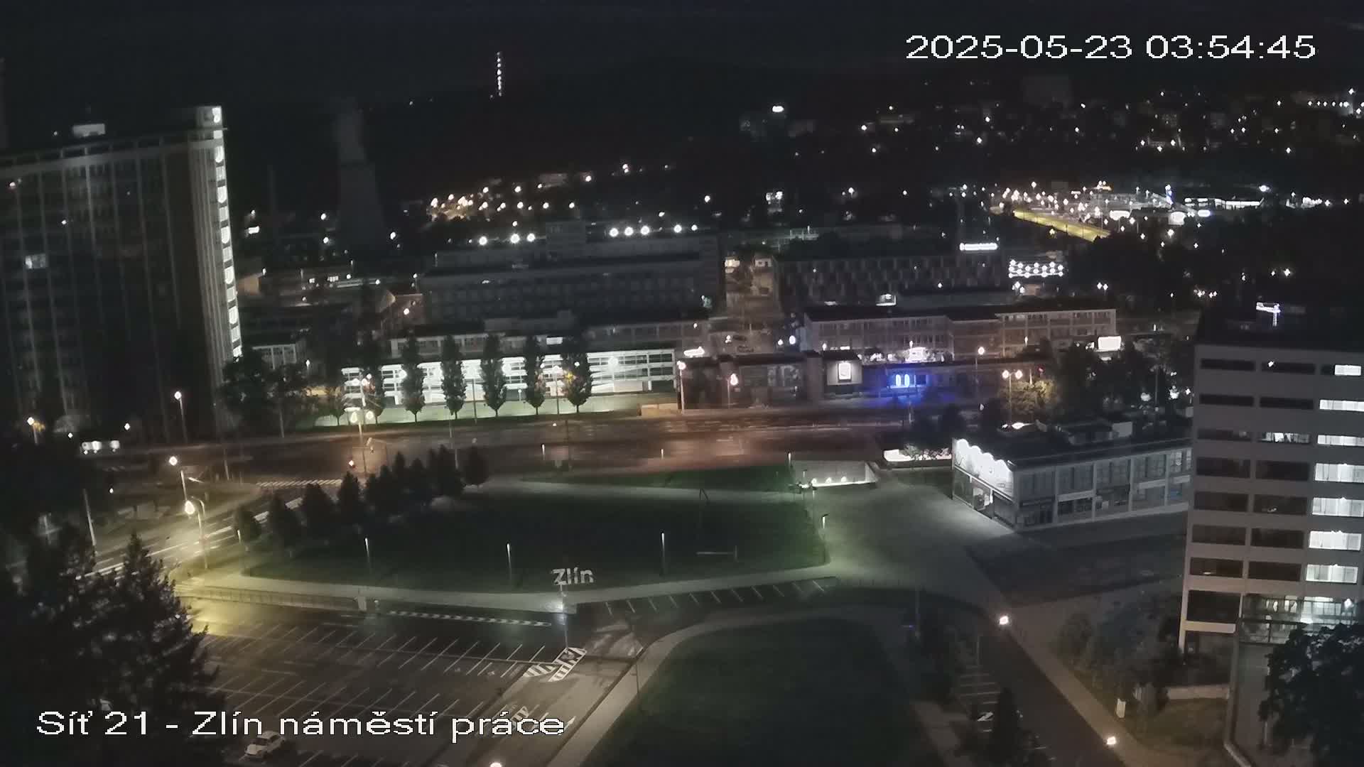 An aerial nighttime view of a city with illuminated buildings and streets, showing a mostly empty parking lot and a park-like area.