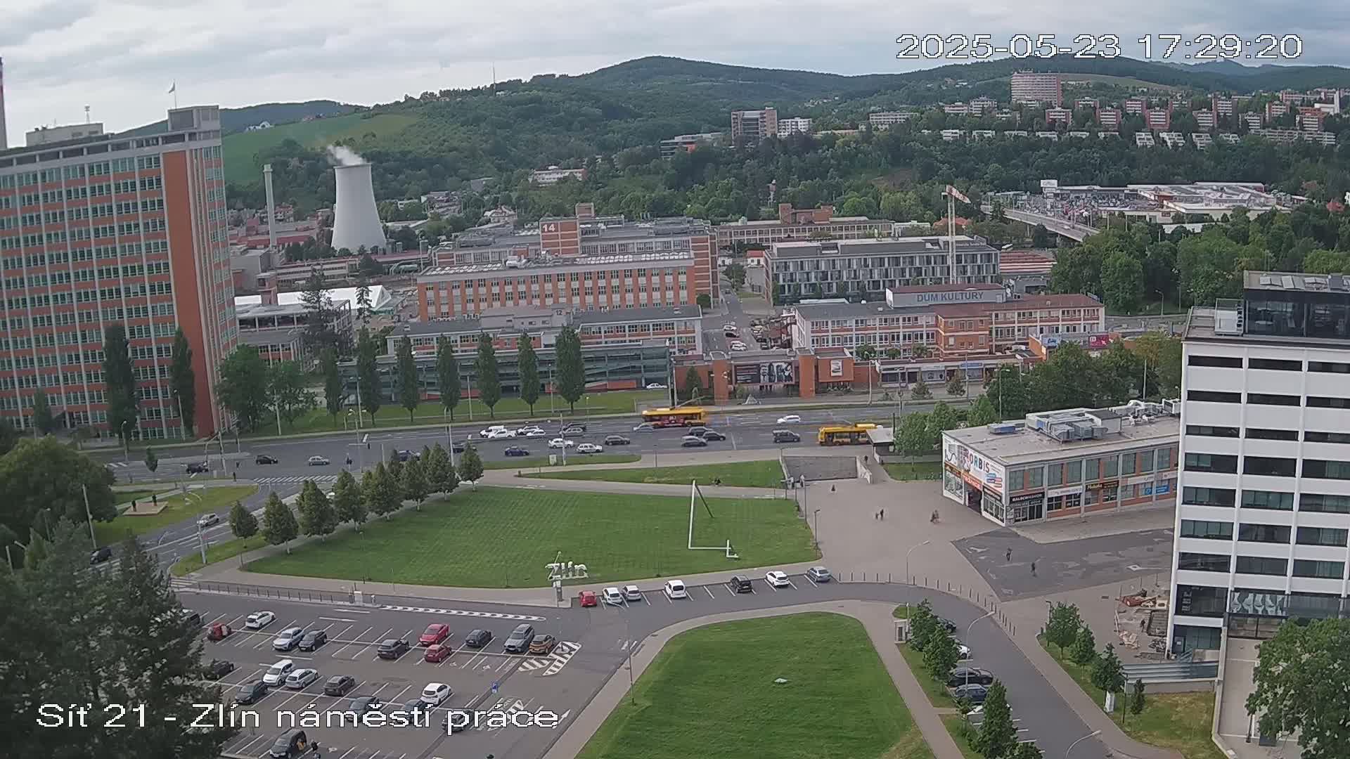 An aerial view of a city on an overcast day, showing a large grassy area with a parking lot and several buildings, including a tall brick building and a large industrial structure with a smokestack, with cars and buses traveling along a road.