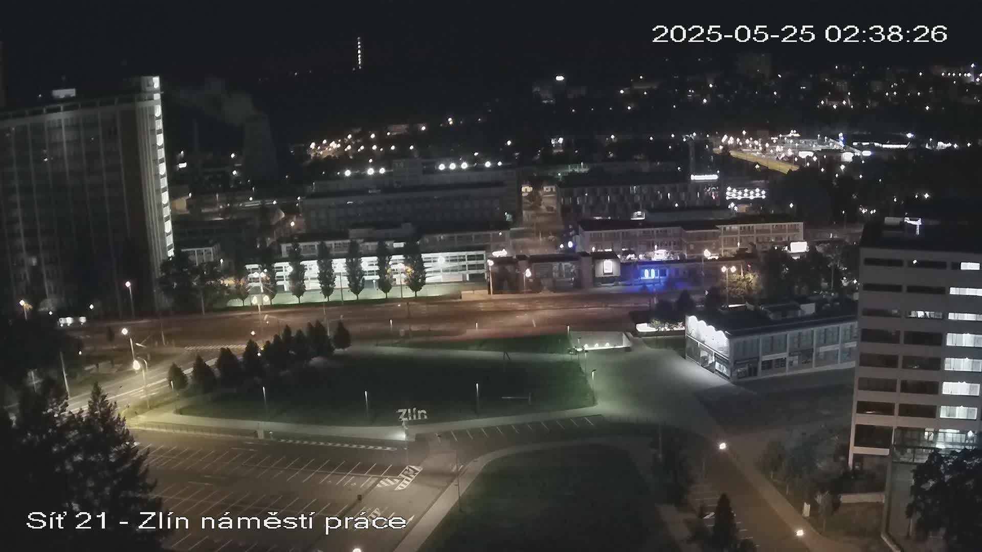 A nighttime aerial view of a city square with several buildings, streets, and parking lots, illuminated by streetlights.