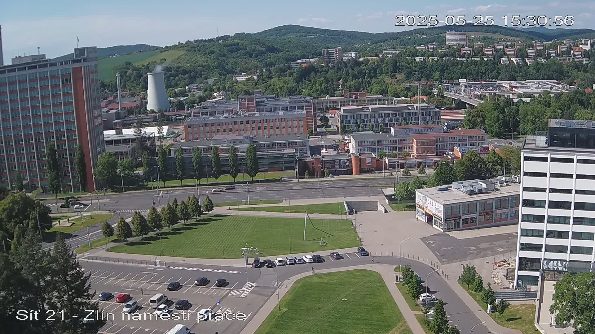An aerial view of a city on a sunny day shows buildings, roads, green spaces, and a parking lot.