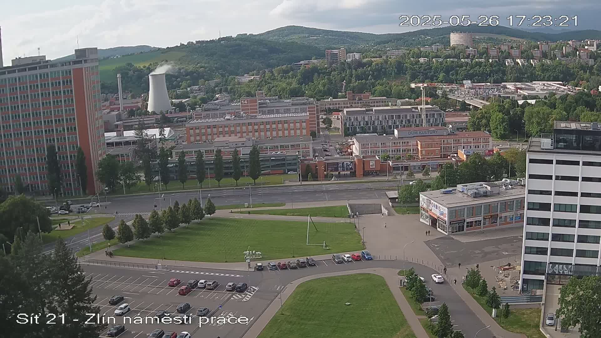 An aerial view of a city scene on a partly cloudy day, showing a mix of buildings, parking lots, and green spaces, with a cooling tower visible in the distance.